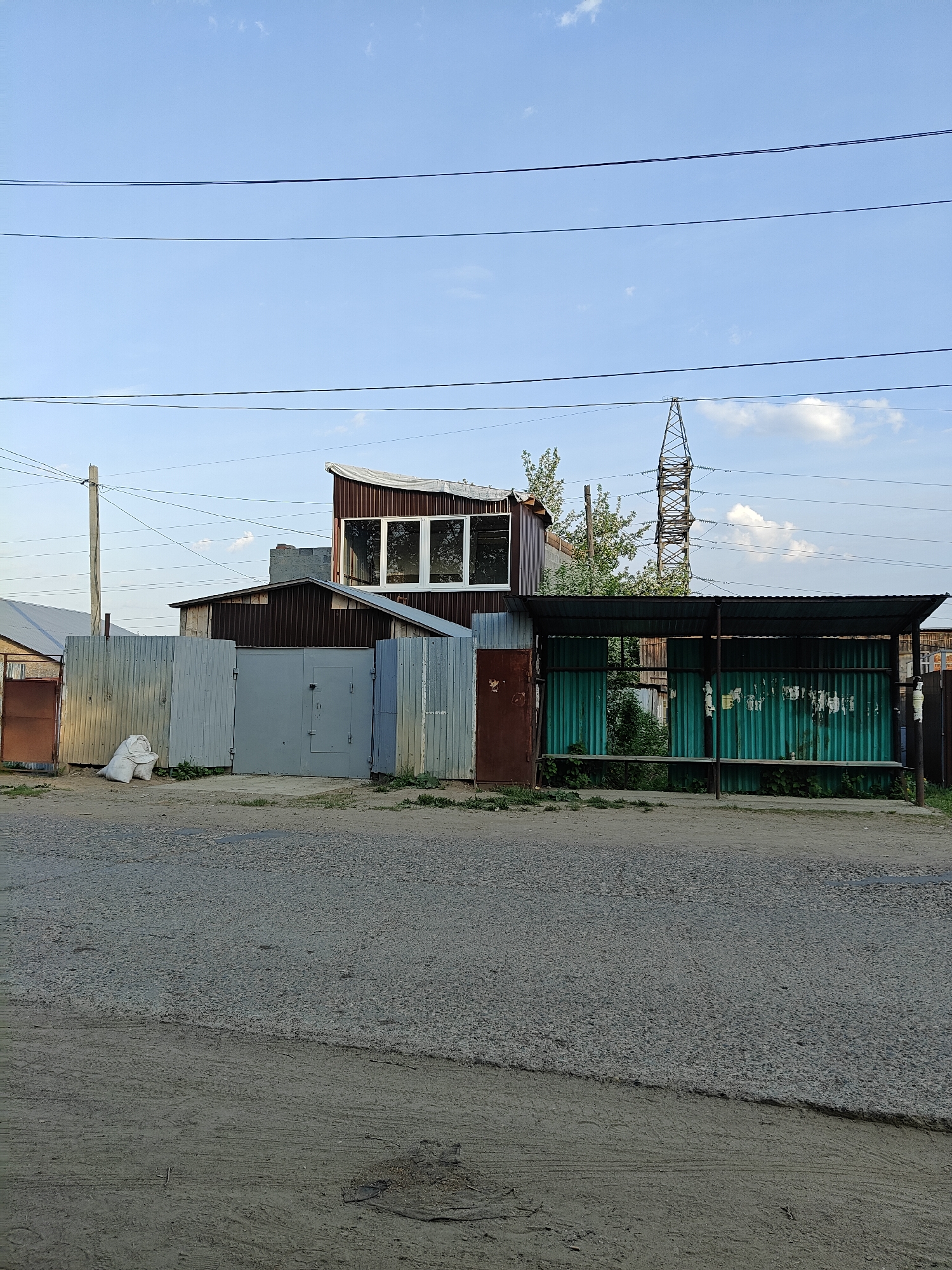 The image shows a street scene in what appears to be a residential area of Chaikovsky, Russia. In the foreground is a dirt and gravel road. Behind the road, there are a series of buildings and structures including a metal fence with a gray gate, a brown building with a second-story structure featuring windows, and a bus stop shelter made of green corrugated metal. An electrical transmission tower is visible in the background. Above, there are utility lines stretching across a sky with some clouds. There are no people visible in the image. There are a few bags by the fence to the left of the gray gate.
