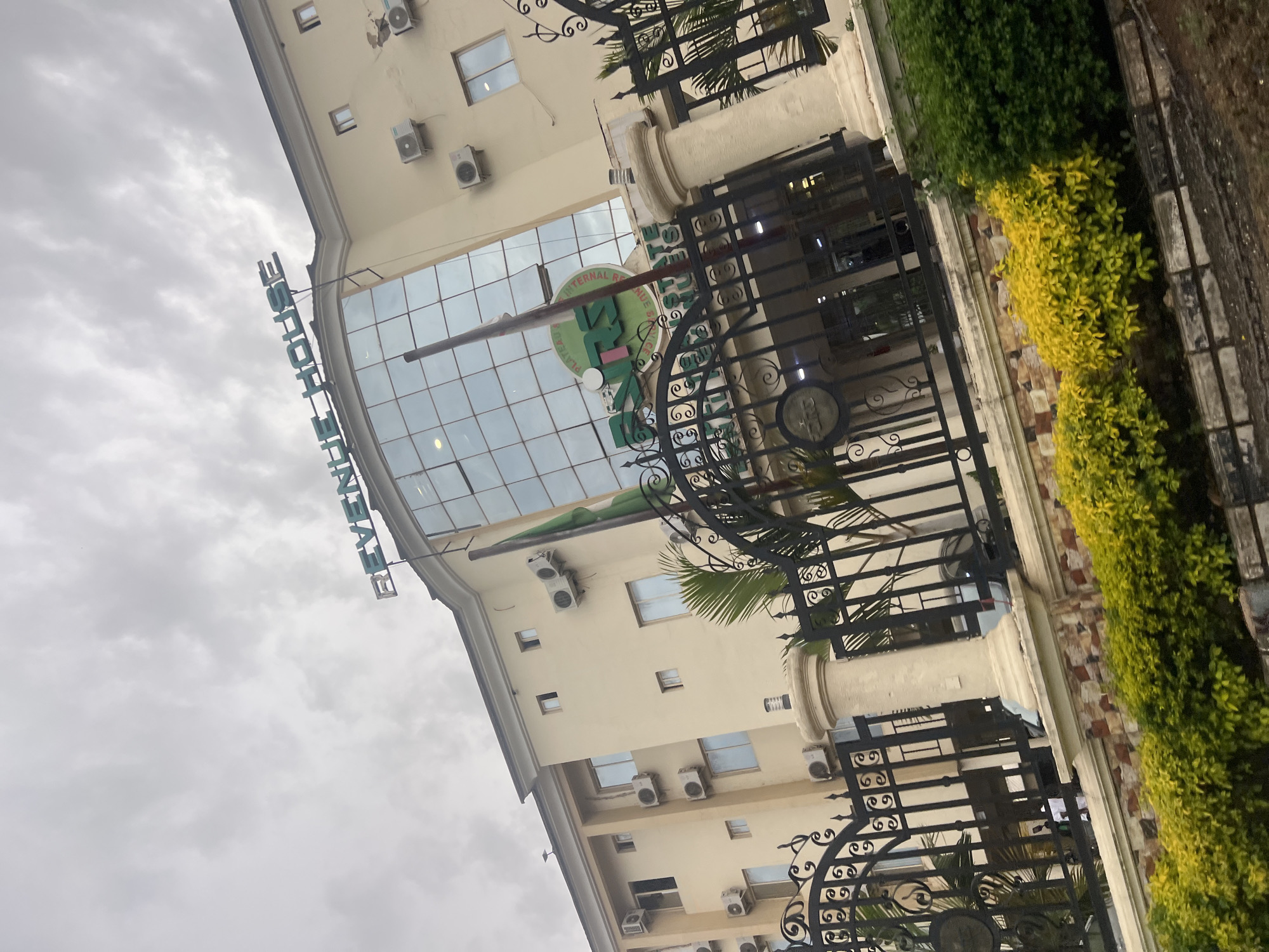 The image shows the Plateau State Internal Revenue Service (Plateau IRS) building, also known as Revenue House, located in Jos, Nigeria. The building is a cream-colored, multi-story structure with a prominent glass facade. A large green circular logo with the inscription "PLATEAU STATE INTERNAL REVENUE SERVICE" and "IRS" is visible on the facade. The building's name, "REVENUE HOUSE," is displayed in large, light blue lettering on the rooftop. The entrance is gated by an ornate black metal fence with decorative pillars. A Nigerian flag is seen flying on the right side of the building. The foreground features a line of yellow and green bushes and a low stone wall. The sky is overcast with clouds.