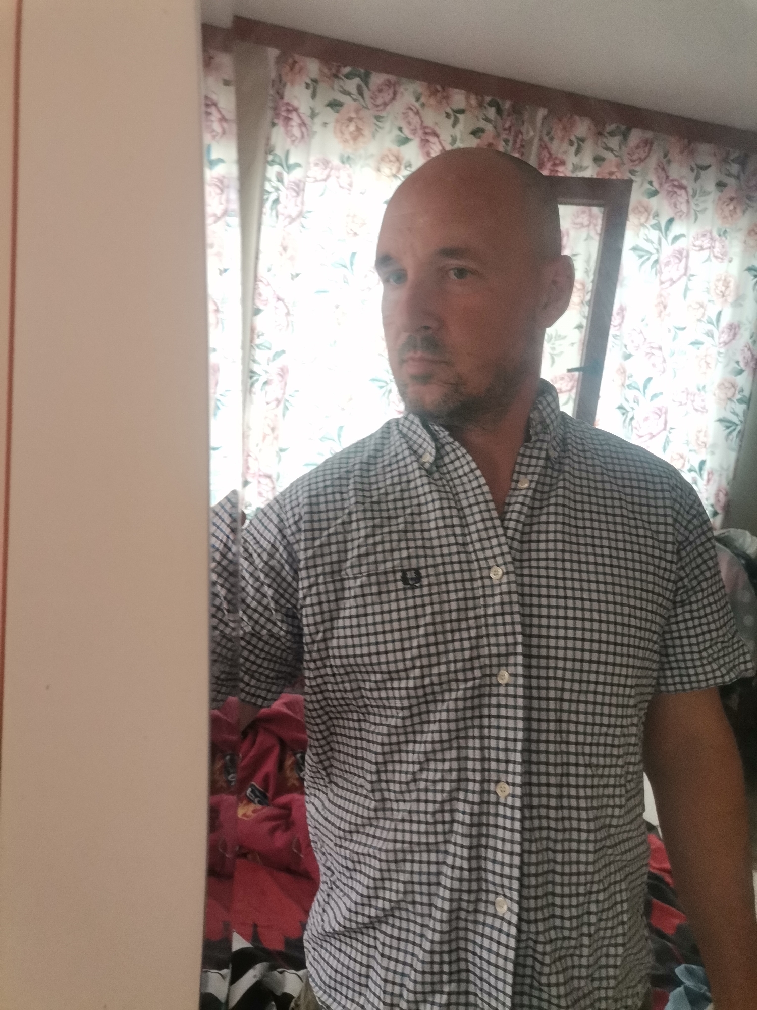 A bald man is standing in front of a mirror. He is wearing a short-sleeved, checkered shirt. Behind him is a window with floral patterned curtains. Part of a door or wall is visible on the left side of the image. The location is in a room in the city of Unknown, Unknown.