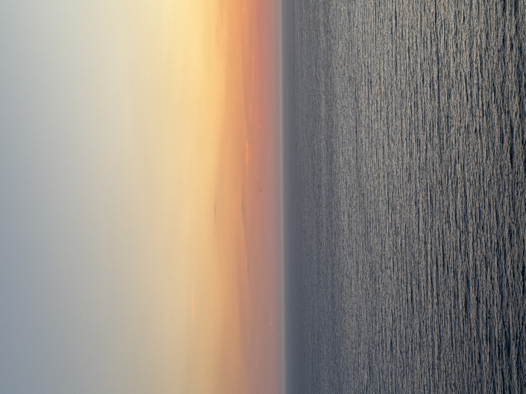 The image depicts a serene seascape at sunset. The sky transitions from a pale blue at the top to a warm gradient of orange and pink near the horizon. A thin band of clouds is visible in the distance, catching the sunset colors. Below the sky lies the calm sea, its surface rippled with gentle waves reflecting the muted tones of the sky. The water is a deep, muted blue-gray. The scene is devoid of any human presence, structures, or distinctive landmarks, suggesting an unobstructed view of the natural environment. The overall impression is one of tranquility and natural beauty, characteristic of coastal landscapes. The location in Glavatičići, Montenegro, implies a possible view of the Adriatic Sea.