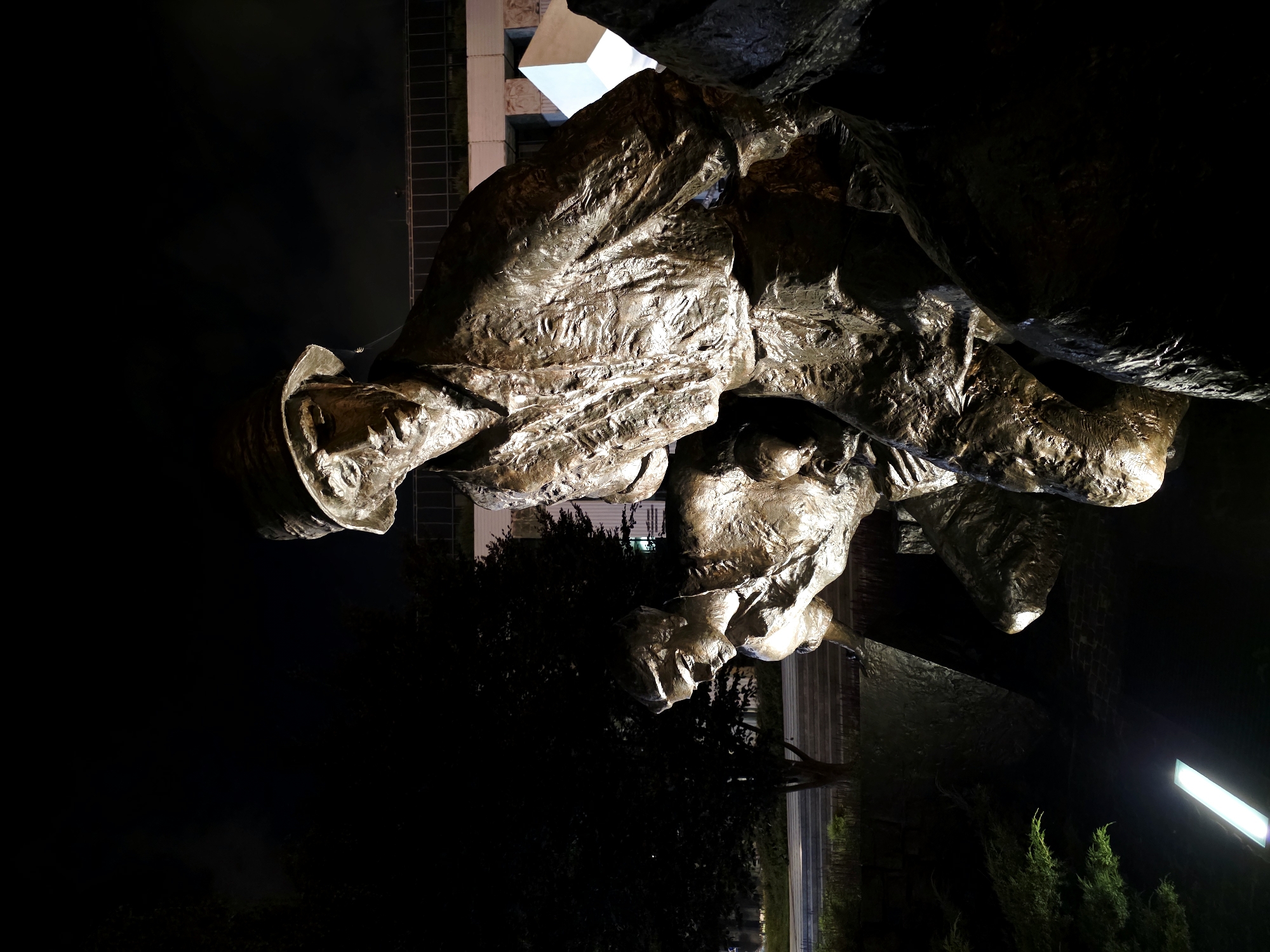The photograph depicts a bronze sculpture of two figures at night, located in Warsaw, Poland. The sculpture appears to be made of weathered bronze and depicts two men in what seem to be military uniforms and hats. The figure in the foreground is larger and more detailed, with his face turned slightly towards the viewer, looking downward. His uniform is textured, suggesting heavy fabric. Behind him and slightly to the left is a second, smaller figure, also in uniform, with his head bowed. The figures are positioned in a way that suggests they are either moving or working together, possibly carrying something or navigating rough terrain, as indicated by the textured, rocky base of the sculpture.
The lighting in the scene is dramatic, with spotlights illuminating the figures from below and to the side, creating strong contrasts and highlighting the metallic texture of the bronze. The background is dark, with the silhouette of trees and a modern building with glass windows visible behind the sculpture. A prominent, angular architectural element, possibly part of the building or another sculpture, is lit up and partially visible on the right side of the image. The overall mood is somber and perhaps commemorative, emphasized by the nighttime setting and the subject matter. The presence of a linear, bright light fixture at the bottom left suggests a well-lit urban or public space. The sculpture is positioned in an outdoor setting, with foliage and what appears to be a paved area visible in the mid-ground.