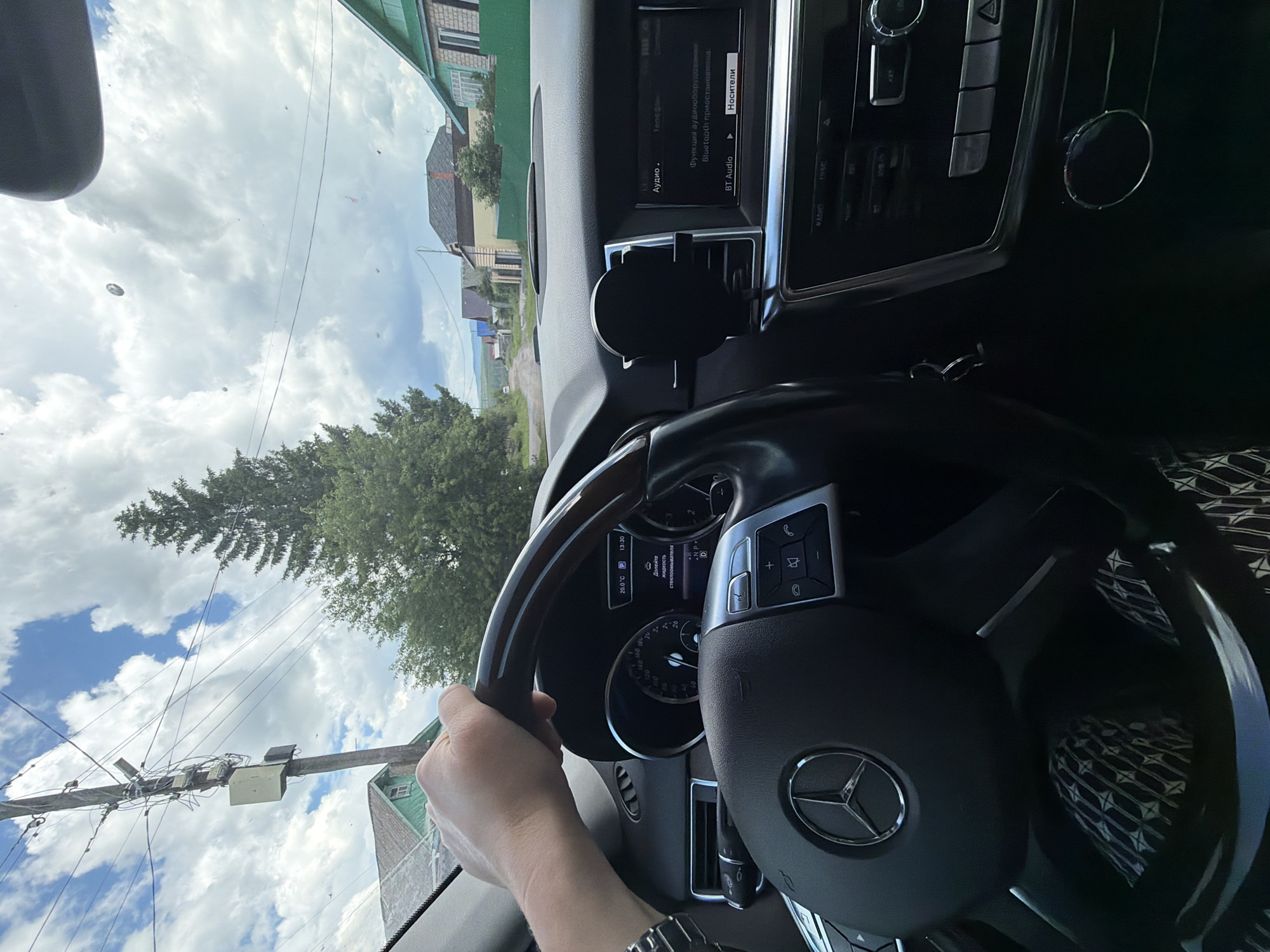 The image is taken from the interior of a Mercedes-Benz vehicle, looking forward through the windshield. A person's hand, wearing a watch, is visible gripping the steering wheel. The dashboard displays a temperature of 20.0 degrees Celsius and the time 13:30. Text on the dashboard indicates "Долито жидкость стеклоомывателя" which translates to "Washer fluid refilled." The center console features a car infotainment system displaying "Аудио" (Audio) and "Телефон" (Telephone) options, along with Bluetooth connectivity information. Outside the car, a rural road is visible, lined with houses and trees, under a cloudy sky. The scene appears to be in Mezhevoy, Russia, given the context provided.