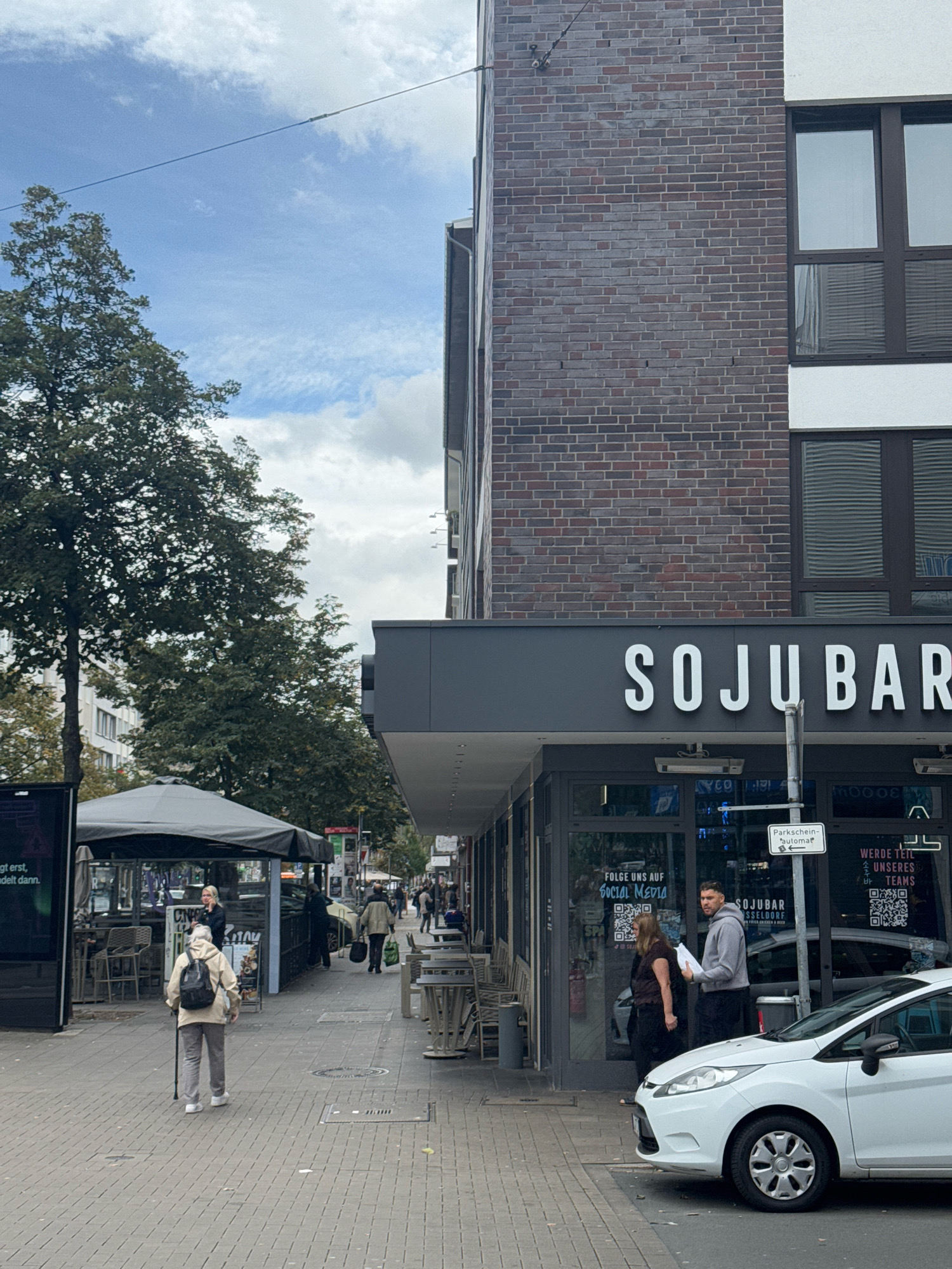 The image depicts a street scene in Dusseldorf, Germany, specifically in front of a business named "SOJU BAR." A woman is walking with a cane on the sidewalk. In the foreground, a white car is parked. Behind the car, a man and a woman are standing near the entrance of the SOJU BAR, engaged in conversation. The bar features large glass windows displaying signage. Further down the street, other pedestrians and businesses are visible. The sky is partly cloudy, and trees line the street.