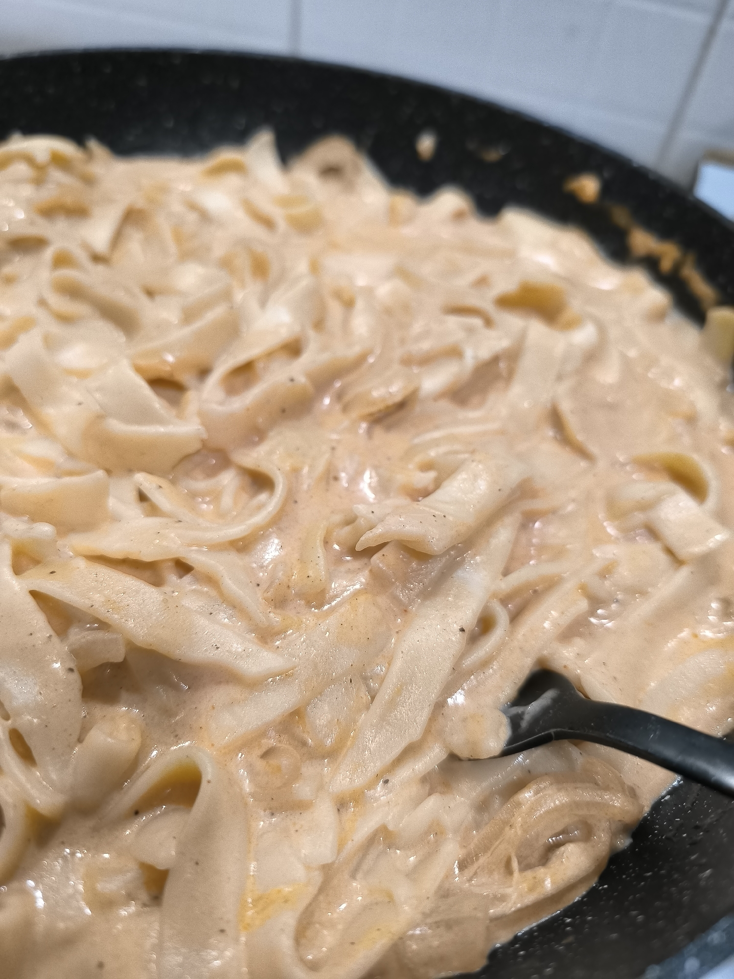 The image shows a close-up of a pan filled with fettuccine pasta in a creamy sauce. A black serving spoon is partially visible, submerged in the pasta. The environment suggests a domestic kitchen setting. No information allows for the location to be identified as Varna, Bulgaria.