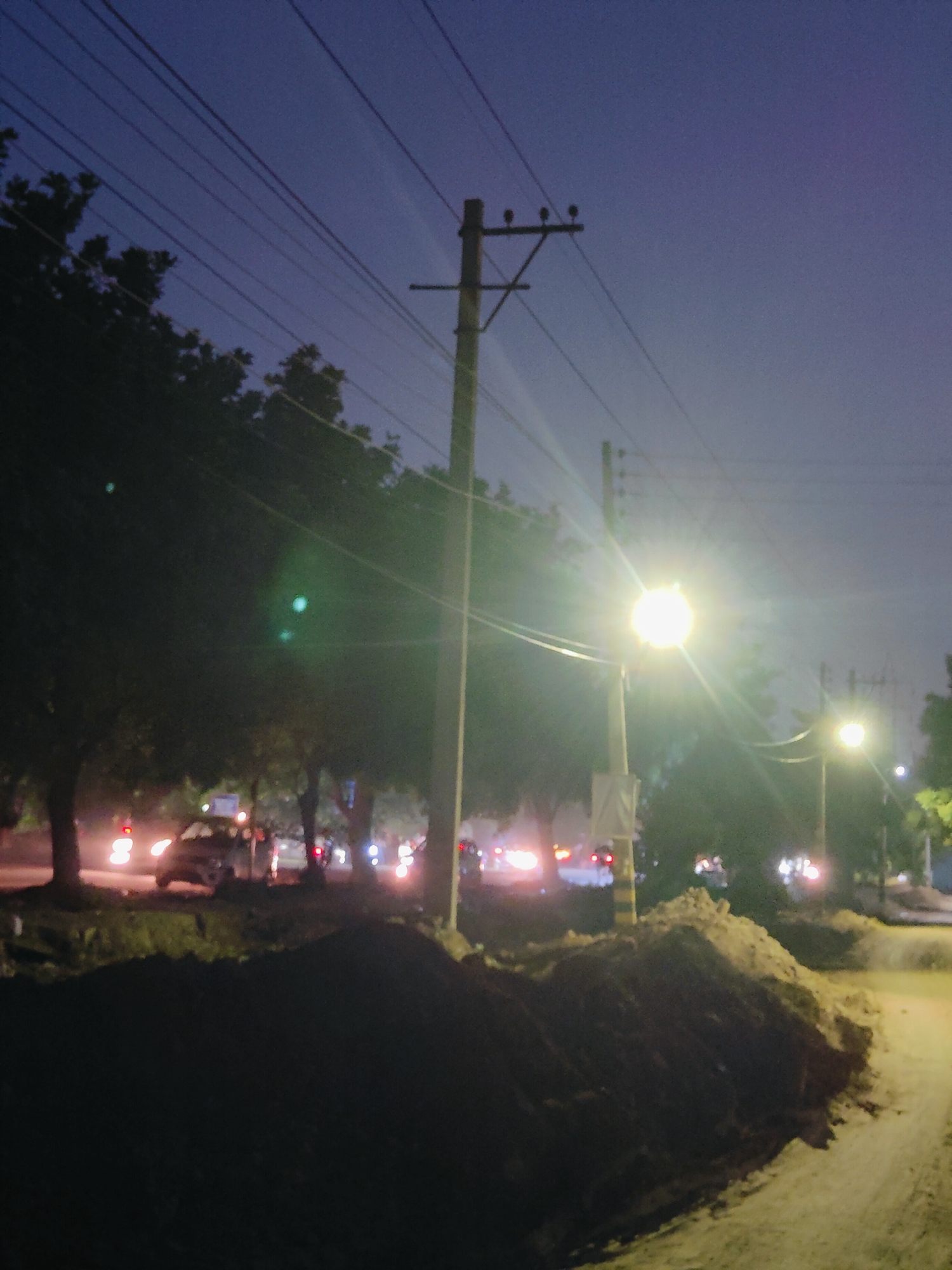 The image depicts a nighttime street scene in Gurgaon, India, characterized by low light conditions and the presence of streetlights and vehicle headlights. Several utility poles with overhead wires are visible, along with the silhouette of trees. In the foreground, there are piles of dirt or construction debris. The background shows a road with moving vehicles, their taillights creating streaks of red light. The sky is a deep blue, suggesting it is either twilight or nighttime. The overall atmosphere is that of an urban environment at night, possibly undergoing some form of roadwork or construction.