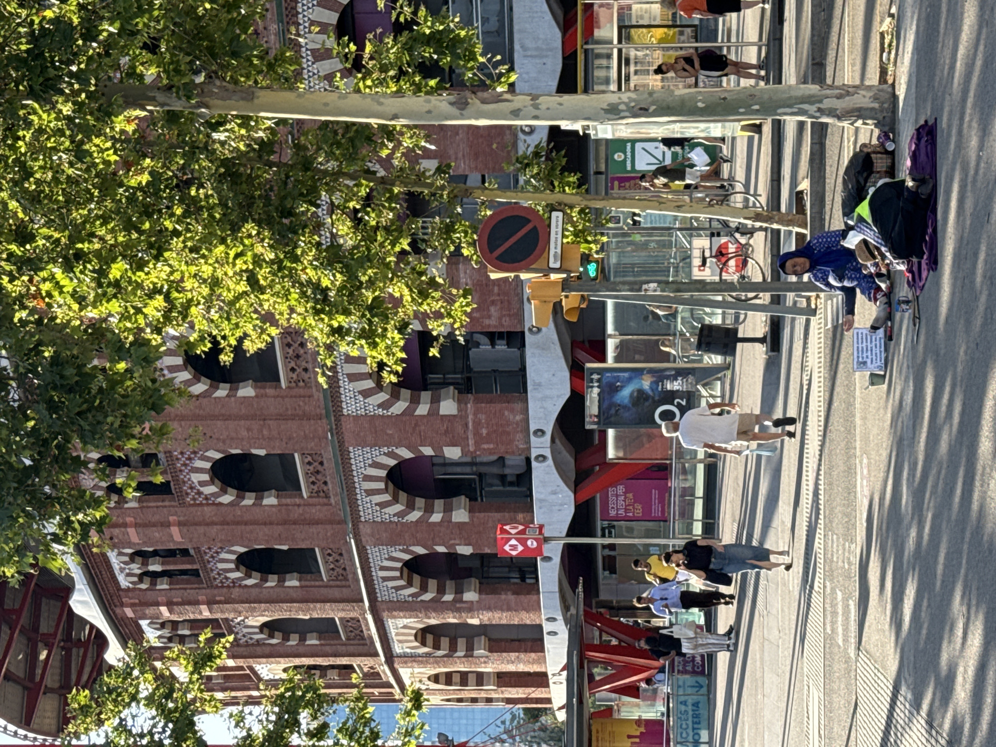 The image depicts a street scene in Barcelona, Spain, likely in the area of the Arenas de Barcelona, a former bullring that has been converted into a shopping center. A large, historic brick building with arched windows and decorative white trim dominates the background, suggesting it is a significant architectural landmark. In the foreground, a woman is sitting on the ground, dressed in a blue headscarf and traditional clothing, appearing to be a homeless individual. She is surrounded by her belongings, including bags and blankets, and has a sign next to her. Other people are walking and milling about in the background, engaged in everyday activities. Trees with lush green foliage line the street, casting dappled shadows on the pavement. Traffic signals and street signs are visible, indicating the urban environment. The overall scene captures a slice of daily life in Barcelona, with a contrast between the bustling city and the plight of a vulnerable individual.