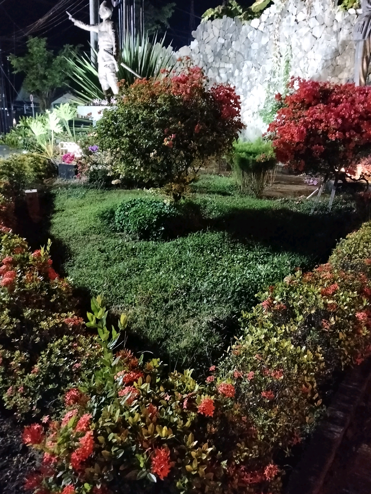 The image shows a monument at night, likely in a public park or square in Purworejo, Indonesia. The monument features a life-sized statue of a person in military-style attire, with one arm extended. The statue is surrounded by lush greenery, including bushes with red flowers and large-leafed plants. In the background, a wall constructed of rough, light-colored stones is visible, with vines growing on it. The scene is illuminated by artificial lighting, which casts shadows and highlights the textures of the stone and foliage. There are no other people visible in the frame, and the overall atmosphere is serene.