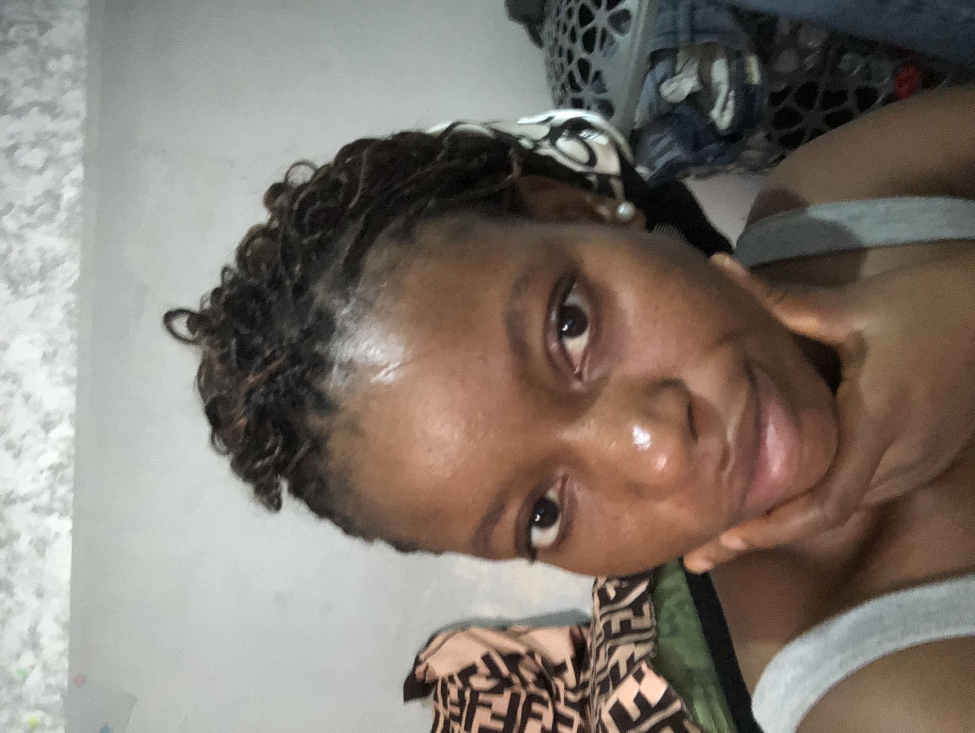 A person with dark skin and short, dark, curly hair is looking directly at the camera with a hand touching their face. They appear to be wearing a gray tank top. The background includes a light gray wall, a patterned blanket or fabric with a geometric design, and a partially visible laundry basket with clothing inside. The location is identified only as Nigeria.