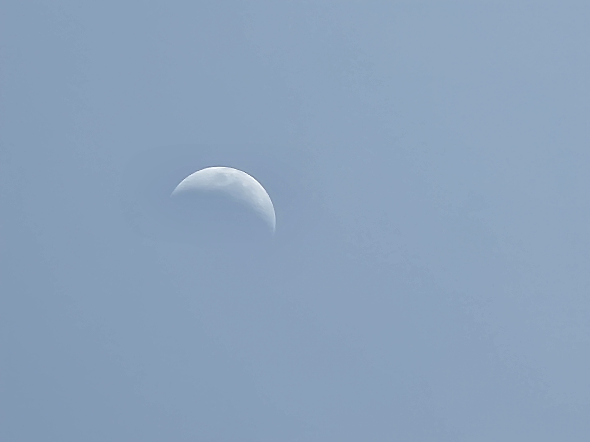 The image is a photograph of a crescent moon in a clear, pale blue sky. The moon is located slightly to the left of the center of the frame and is illuminated from the right. The sky is a uniform color with no visible clouds or other celestial bodies. There are no people, objects, or any identifiable landmarks in the image. The photograph appears to be taken during the daytime, as indicated by the bright sky. There is no information in the image that would allow for the identification of its location in Quarteira, Portugal.