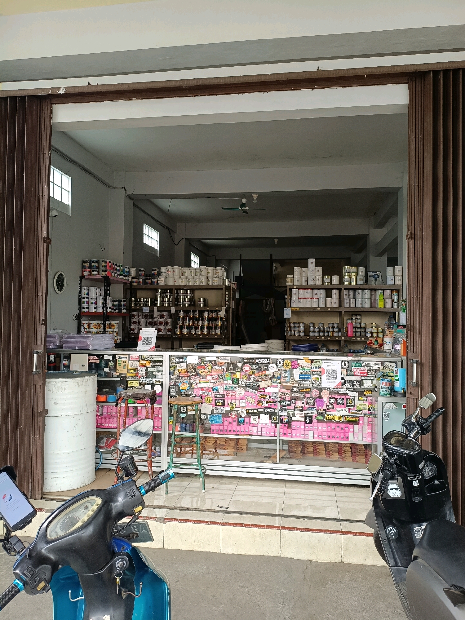 This image depicts the storefront of a shop in Sewon, Indonesia. The shop appears to be an establishment selling various products, possibly paints, chemicals, or craft supplies, given the presence of numerous cans and containers of different sizes on shelves and in display cases. The storefront is open, with a metal roll-up door partially retracted on the right side and a similar door on the left side, also partially retracted.
In the foreground, two motorcycles are parked. The motorcycle on the left is a teal scooter with a phone mount attached to the handlebars, displaying a white screen. The motorcycle on the right is a black scooter.
The interior of the shop is visible, showing shelves filled with merchandise. A glass display counter is prominent in the center, adorned with a large number of colorful stickers. Behind the counter, more shelves are stocked with products. A QR code is visible on the counter.
The environment appears to be an urban or semi-urban setting, with tiled flooring in front of the shop. The shop walls are painted in light colors. Natural light enters from the open doorway, and some artificial lighting fixtures are visible on the ceiling. The overall impression is that of a small business open for trade.
