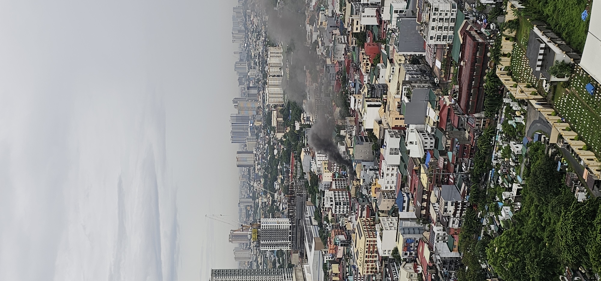 The image is a high-angle, panoramic view of a dense urban landscape in Makati, Philippines, under an overcast sky. In the foreground, a lush green cemetery with numerous white tombstones and mausoleums is visible, bordered by a beige, arched wall. Beyond the cemetery, a densely populated residential and commercial area is spread out, characterized by multi-story buildings with varied roofing colors, including red, green, and gray. Construction cranes and unfinished buildings indicate ongoing development. In the mid-ground and background, a sprawling cityscape of high-rise buildings stretches towards the horizon, interspersed with trees. A prominent feature in the mid-ground is a thick plume of dark smoke rising from a cluster of buildings, suggesting a fire or industrial activity. No people are clearly discernible in the image. The overall impression is of a busy, developing urban environment marked by recent or ongoing events.