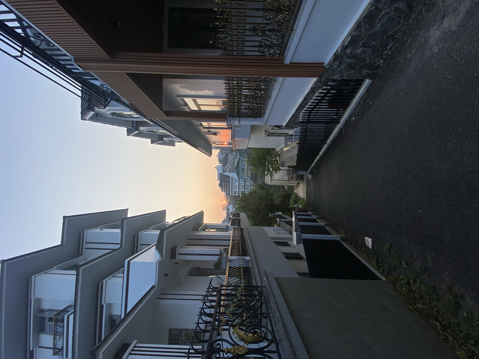 Captured at dusk, the image showcases a narrow street in Lâm Viên - Đà Lạt, Vietnam. The architecture on either side of the street features modern, multi-story buildings with projecting balconies. One side displays ornate wrought-iron railings with gold accents, contrasting with the sleek, geometric design of the buildings on the opposite side, which have prominent white and gray balconies. The street is paved with asphalt, and the fading light of the sunset casts a warm glow between the buildings. In the distance, the city skyline is visible, punctuated by a few taller structures and the tops of trees. No people are present.