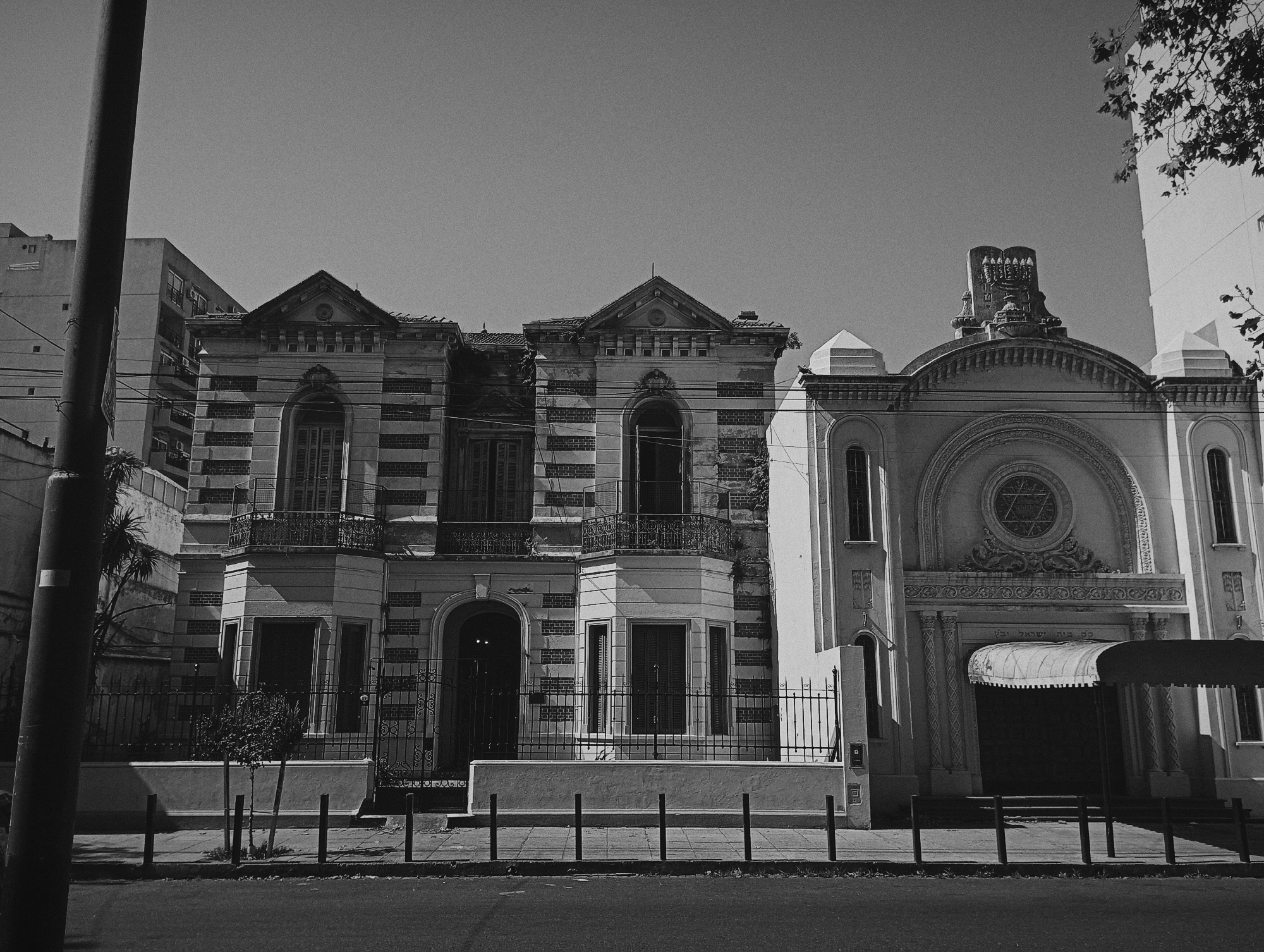 A black and white street scene captures a row of buildings under a clear sky during daylight hours. The image is taken from a slightly low angle, looking up at the structures.
On the far left, a tall, dark utility pole stands prominently, partially obscuring a modern, multi-story building with visible windows and balconies in the background.
The central part of the image features two ornate, older buildings, possibly a large mansion or institutional structure. These buildings share a distinctive architectural style characterized by alternating horizontal bands of light and dark material, likely stone or brick. They feature several arched windows, some with closed shutters, and balconies with decorative iron railings on the upper floors. The roofs are gabled and appear somewhat weathered, with some signs of age or disrepair. A low concrete wall and a decorative black metal fence separate these buildings from the sidewalk. In front of them, a small, leafy plant and another small tree are visible behind the fence.
To the right of these older buildings stands a larger, more imposing structure that is distinctly a synagogue. Its facade is a lighter color, likely white or light gray, with a grand arched entrance. Above the main archway, an intricate circular window contains a prominent Star of David symbol. Below this circular window and above the entrance, faint Hebrew text is visible. Further above, on the roofline, an ornate sculpture depicts the Tablets of the Ten Commandments, topped with a menorah, clearly identifying the building's religious purpose. A retractable awning extends over the synagogue's main entrance. On either side of the central synagogue facade, there are simpler, recessed sections with narrow, arched windows.
In the immediate foreground, a paved street or road is visible, followed by a sidewalk. Along the curb, several black, vertical bollards are evenly spaced. The overall lighting suggests a bright, sunny day, with distinct shadows cast by the buildings and street furniture. Tree branches are visible in the upper right corner of the frame, hinting at larger trees outside the immediate view. The black and white photography lends a timeless or historical quality to the scene.