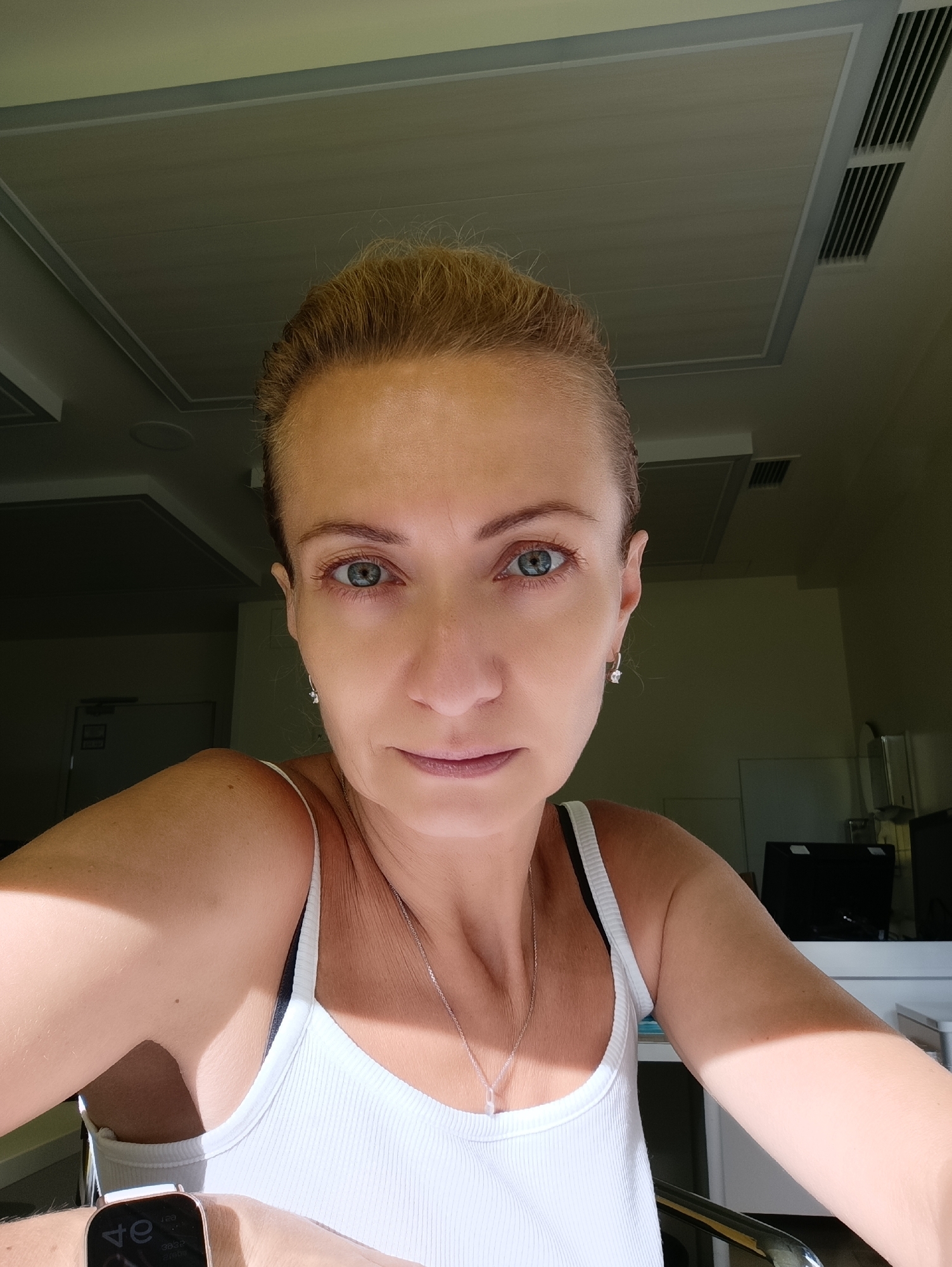 The image shows a medium shot of a woman with blonde hair pulled back. She is wearing a white ribbed tank top with a black bra underneath and a silver necklace. She is looking directly at the camera with a neutral expression. Sunlight is hitting her face and shoulder from the left. The background appears to be an indoor setting with a drop ceiling and air vents. There is a desk with a computer monitor visible in the background. The woman is wearing a smartwatch on her left wrist. There is no indication that this image was taken in Moscow, Russia.