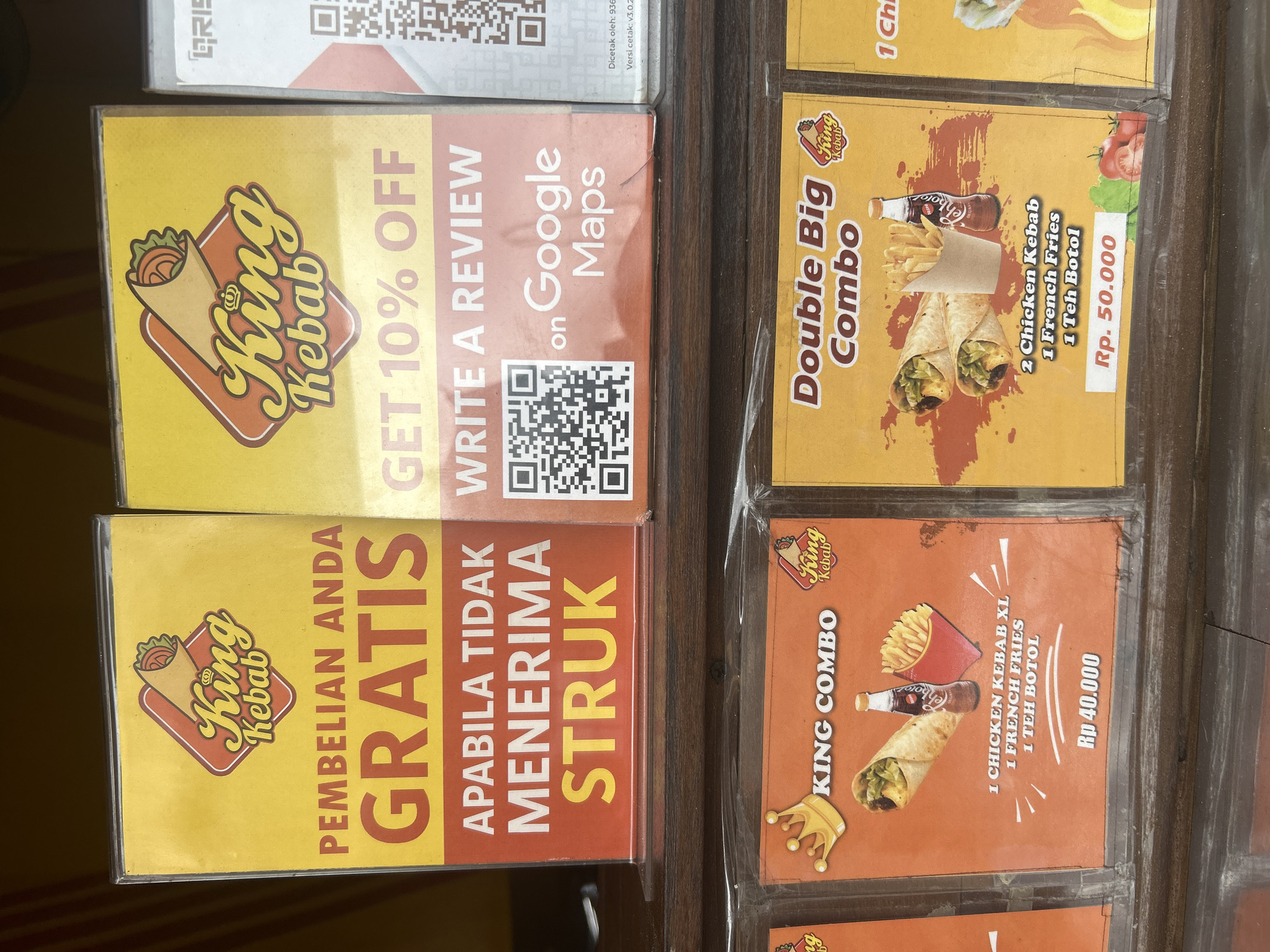 The image displays a close-up of several laminated promotional flyers for "King Kebab" restaurant. The flyers are arranged in a grid, with some showing food items and others offering discounts or promotions.
The top left flyer is mostly obscured, but shows a QR code and text that appears to be in Indonesian. Below it, a yellow flyer announces "PEMBELIAN ANDA GRATIS" (Your Purchase is FREE) and "APABILA TIDAK MENERIMA STRUK" (If you don't receive a receipt), indicating a promotion related to receipts. It also features the "King Kebab" logo, which is a stylized drawing of a kebab.
To the right of these, a yellow flyer advertises a "Double Big Combo" for Rp. 50,000, which includes "2 Chicken Kebab, 1 French Fries, 1 Teh Botol" (1 Chicken Kebab, 1 French Fries, 1 Bottled Tea). An image of the combo, featuring two kebabs, french fries, and a bottle of what looks like soda, is prominently displayed.
Below the "GRATIS" flyer, another yellow flyer promotes "GET 10% OFF WRITE A REVIEW on Google Maps" and displays a QR code for Google Maps.
The bottom right flyer, with an orange background, advertises a "KING COMBO" for Rp 40,000, featuring "1 CHICKEN KEBAB XL, 1 FRENCH FRIES, 1 TEH BOTOL" (1 XL Chicken Kebab, 1 French Fries, 1 Bottled Tea), along with an image of the combo.
The overall scene suggests a food establishment, likely a kiosk or small restaurant, where these promotional materials are displayed to attract customers. The Indonesian text and currency (Rp) confirm a location in Indonesia, specifically Denpasar as indicated in the prompt. The lighting suggests it could be daytime. There are no people visible in the image, only the promotional materials and a wooden surface, possibly a counter or table, on which they are placed.