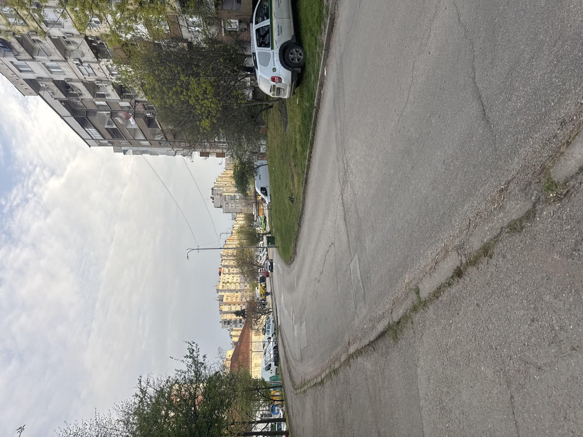 The image depicts a street scene in Montana, Bulgaria, characterized by aging infrastructure and a typical urban environment. Tall apartment buildings, indicative of Soviet-era construction, line the street, suggesting a residential area. The road is paved but shows signs of wear and tear. Vehicles, including a white SUV with a "John Deere" logo, are parked along the roadside, which is bordered by a narrow strip of grass. Trees with budding leaves are present, hinting at springtime. The sky is overcast, contributing to a somewhat bleak atmosphere. The overall impression is that of a working-class neighborhood in a post-socialist city.
