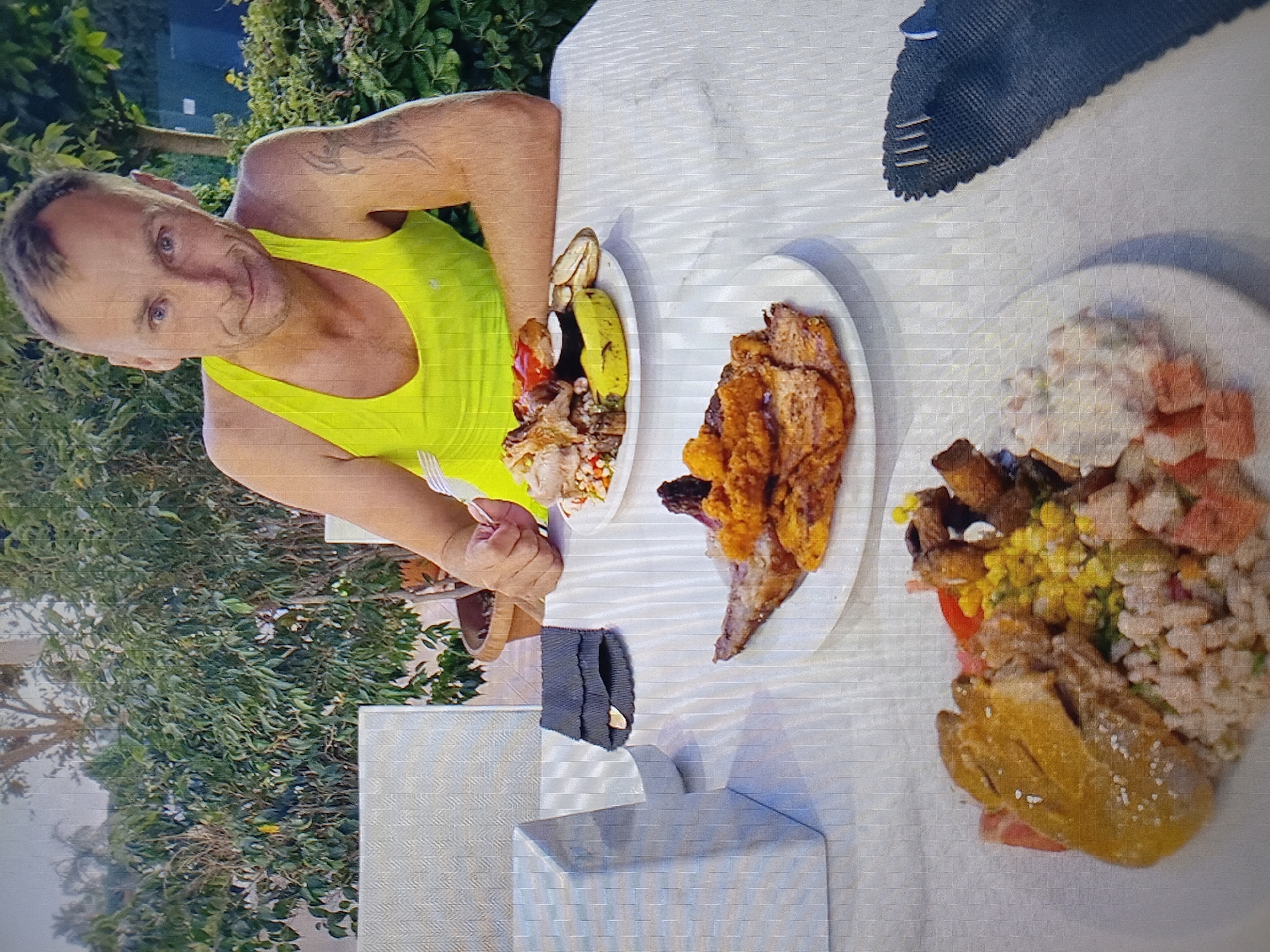 A man wearing a neon yellow tank top sits at a white table laden with food, in an outdoor setting. He is looking directly at the camera with a neutral expression. In front of him are two plates of food. The plate closest to him contains a variety of meats, vegetables, and grains. Further away, another plate holds more meat and vegetables, including what appears to be grilled chicken and some form of salad. A fork rests on the table near the man's hand. The background shows lush green foliage and trees. The image does not provide any specific details or context that would place the scene in Tiraspol, Moldova.