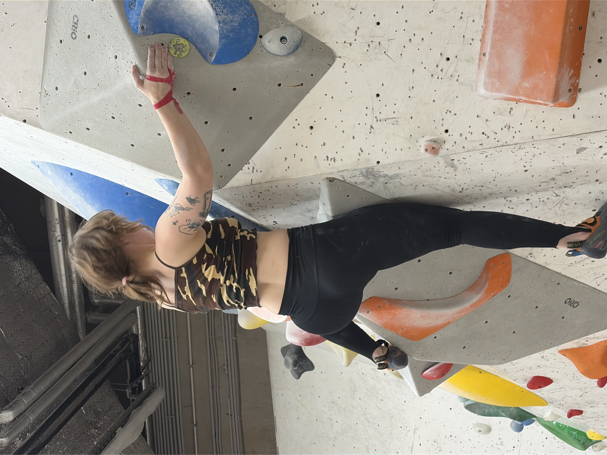 An adult female with brown hair tied back in a ponytail is rock climbing indoors. She is wearing a camouflage print crop top and black leggings, with red tape around her left wrist. Her right hand is on a climbing hold on a large, textured white wall. Various colored climbing holds are visible on the wall, including blue, yellow, orange, and red. The environment appears to be a climbing gym, with pipes and insulation visible on the ceiling. The scene is well-lit, with the primary light source coming from above. The image was taken in Moscow, Russia.