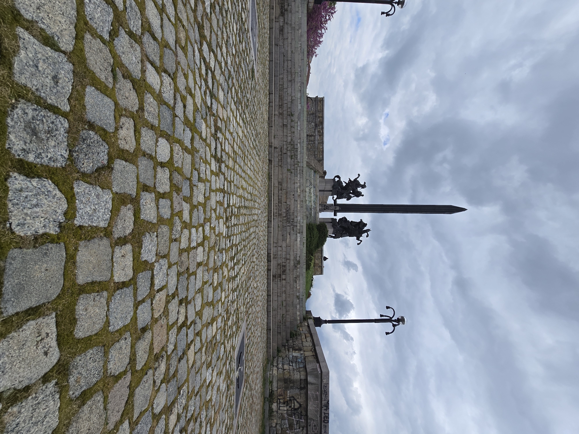 A medium shot, rotated counter-clockwise, depicts an outdoor setting with gray cobblestone paving in the foreground, leading towards a monument featuring bronze equestrian sculptures and a tall obelisk against a cloudy sky. To the right, tiered stone steps ascend towards the monument, flanked by vintage-style lampposts. Graffiti is visible on a low retaining wall near the cobblestone. The location is in the city of Unknown, Unknown.