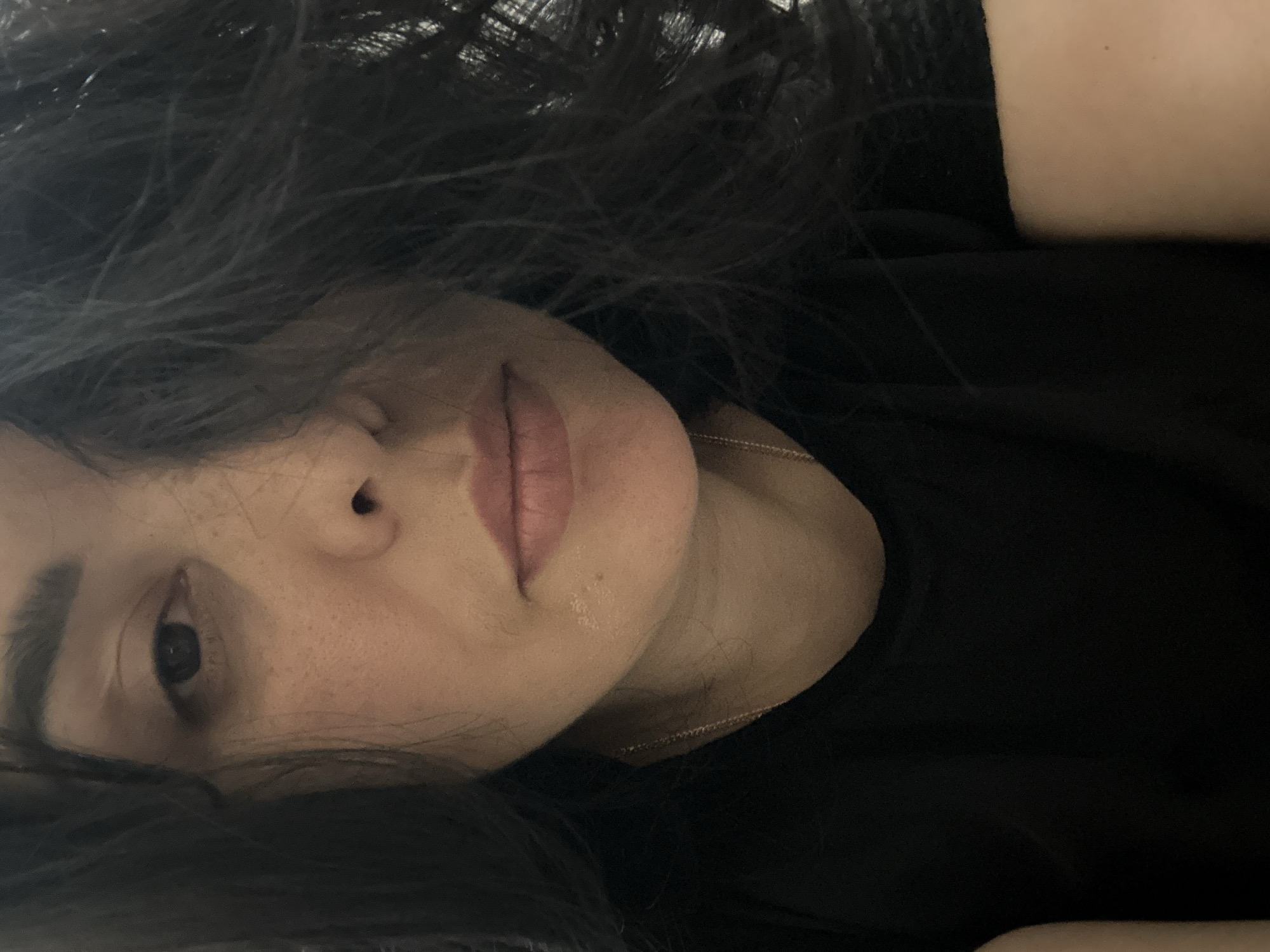 A close-up, horizontally oriented, angled shot focuses on the face of a light-skinned individual with dark hair. The subject is wearing a black shirt and a delicate gold chain necklace. One eye is visible, and the nose and mouth are prominent features. The individual appears to be lying down. The location is unknown.