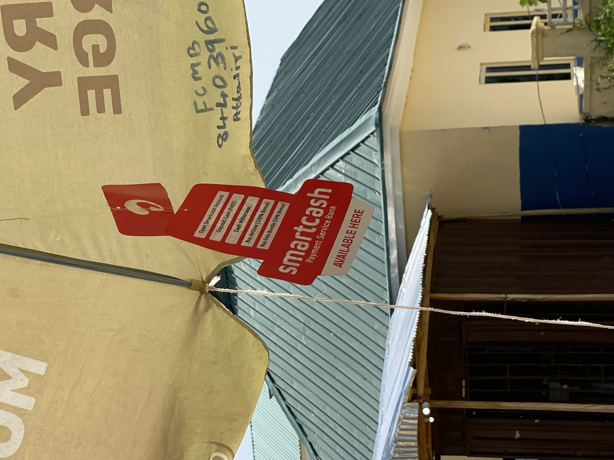 The image shows an outdoor scene, likely in Jalingo, Nigeria, on a sunny day. The main subject is a light-colored umbrella, possibly for a stall, which has several red tags attached to it. These tags advertise services offered by "smartcash Payment Service Bank," including opening an account, depositing cash, withdrawing cash, buying airtime with a bonus, and buying data bundles. Text on the umbrella itself is partially visible, with the letters "ҮЯ" and "อ" on the left side.
To the right of the umbrella, a green corrugated metal roof is visible, suggesting a building or structure. Further to the right, a building with cream-colored walls and dark-framed windows is seen. The top portion of this building features two small, rectangular windows. A blue section is painted on the lower part of this building.
Handwritten text on the umbrella reads "FCMB 84403960 Attasiri," which likely refers to a bank and account or contact information. A thin white string is stretched across the lower part of the image, appearing to be part of a makeshift setup. The overall impression is a bustling market or roadside stall offering financial services.