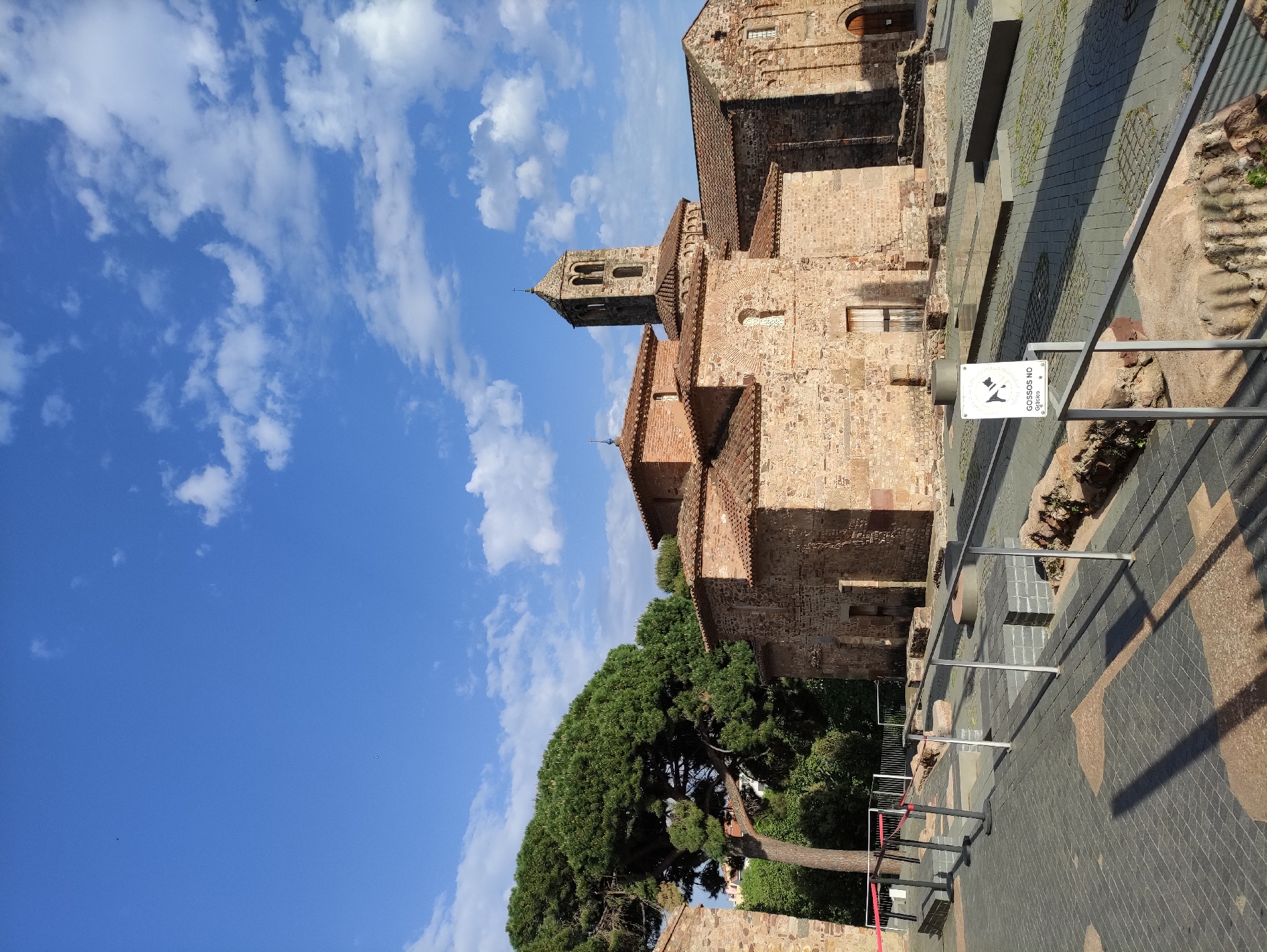 This image captures a vibrant daytime scene of a historical site, likely an archaeological park or a monastery complex, bathed in bright sunlight under a clear blue sky with scattered white clouds. The setting is characterized by ancient stone architecture and preserved ruins.
The prominent subject is a large, sturdy stone building, typical of Romanesque or Gothic architecture, featuring a distinctive bell tower (campanile) with arched openings. The building is constructed from rustic, reddish-brown stone blocks and boasts terracotta tiled roofs. Further back and to the right, another section of a similar stone building is visible, with a small window and a dark arched doorway.
In the foreground, to the left, a large, mature pine tree with a dense, umbrella-shaped canopy adds a natural element to the historical landscape. Its dark green foliage contrasts with the clear blue sky.
The ground features paved areas with varying patterns of grey and light brown paving stones. Sections of excavated ruins or ancient foundations, composed of rough stone, are visible and protected by modern metal railings. These railings guide visitors through the site, separating the pathways from the historical remnants.
A white sign, mounted on a metal pole, is clearly visible in the mid-ground. It displays a black icon of a dog crossed out, with the text "GOSSOS NO" and "Gràcies" underneath. This Catalan text translates to "No Dogs" and "Thank you," indicating a local rule for the preservation of the site.
The strong shadows cast by the buildings and railings suggest a time of day with high sun, likely mid-morning or early afternoon. The overall atmosphere is one of historical significance, peace, and good weather.