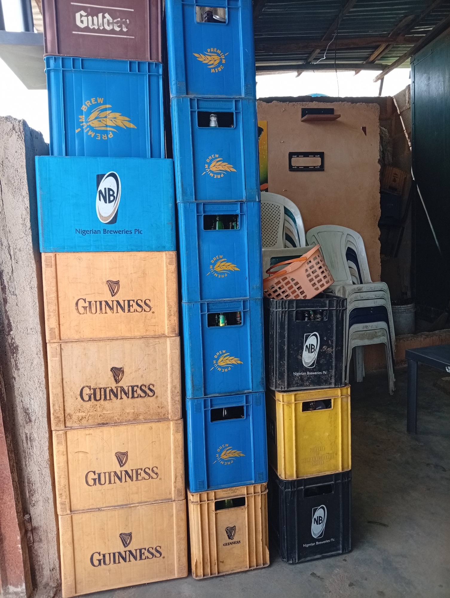The image shows a stack of beer crates in an outdoor setting, likely a bar or storage area in Jos, Nigeria. On the left, a stack of Guinness crates in shades of orange and tan is visible, bearing the Guinness logo. Next to them is a tall stack of blue "Premium Brew" crates, some with bottles still inside. To the right, shorter stacks of black, yellow, and blue crates with the "NB" logo are visible. Stacked plastic chairs and a basket are in the background, along with a glimpse of a building interior with shelves. The scene suggests a typical Nigerian establishment where beer is sold and stored.