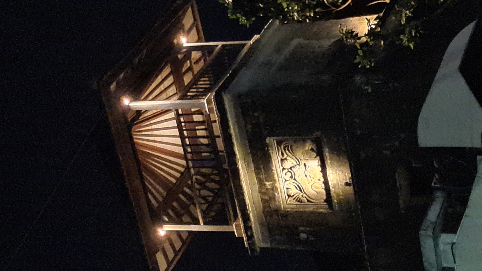 The image captures a dimly lit exterior scene, possibly at night, showcasing elements of traditional architecture. The primary subjects are portions of a building with intricate design. On the left, a wooden roof structure with angled beams is illuminated by a warm light source. Supporting this roof are white pillars. Adjacent to this, a darker wall of the building features a carved relief panel, also lit, displaying ornate patterns that suggest cultural or historical significance. A tree with visible leaves is present in the upper right, its branches casting shadows. To the far right, a lighter, angular architectural element is partially visible, contrasting with the darker tones of the main structure. The scene appears to be outdoors, given the presence of foliage and the overall lighting, which hints at an artificial light source. The atmosphere is quiet and perhaps serene, with the lighting drawing attention to the architectural details. The context of Denpasar, Indonesia, suggests this could be a temple, a traditional house, or a historical monument, common architectural styles in the region. No people are visible, nor is any text. The time of day is clearly night or very late evening due to the darkness and artificial lighting.