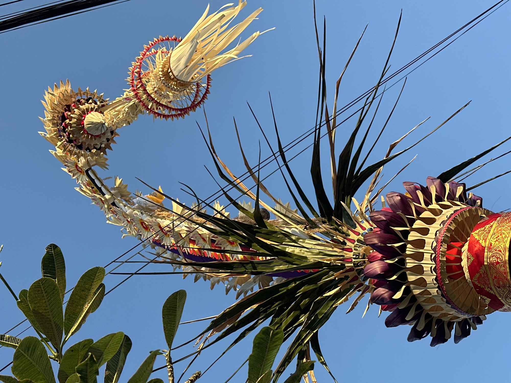 A decorative Balinese Janur Kuning, a traditional offering made from young coconut leaves, is displayed against a clear blue sky. The elaborate structure is adorned with woven leaves, dried flowers, and colorful ribbons of red, purple, and yellow. The base of the offering is richly decorated with red and gold fabric featuring intricate patterns. Two sets of black wires, likely power lines, are visible against the sky, intersecting the frame. The lower left portion of the image shows large green leaves from a nearby tree, possibly a Frangipani, adding a natural element to the scene. The photo appears to have been taken in the city of North Kuta, Indonesia, given the context of such offerings being common in Balinese culture.