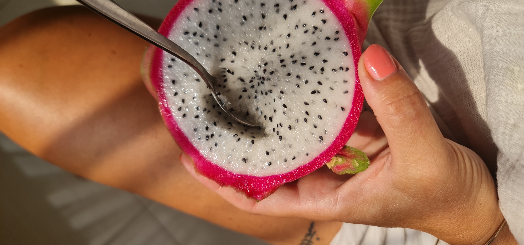 A close-up shot depicts a person holding a halved dragon fruit with a spoon partially inserted into the white flesh dotted with black seeds. The dragon fruit has a vibrant pink rind and a green stem. The person's hand, with coral-colored nail polish, is holding the fruit against a backdrop of what appears to be a light-colored garment. Part of the person's arm is also visible. The setting's location in Đà Nẵng, Vietnam, is not evident from the visual information provided.