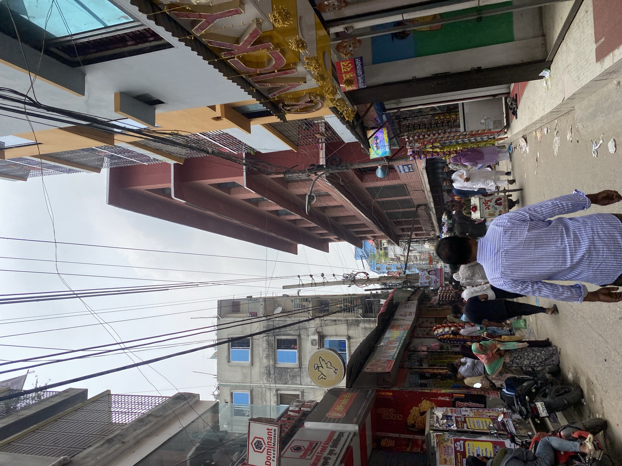 The image shows a street scene in Dhaka, Bangladesh. The view is along a narrow street bordered by buildings of varied height and architectural style. Power lines crisscross above the street. On the right side of the street, people are walking. Some are wearing traditional South Asian clothing. Several motorcycles are parked along the curb. Storefronts feature signage in both English and Bengali. Signs for "Dominant Food Museum" and "Crispy Chicken" are visible. A taller building in the background has multiple windows. The sky is overcast. The photo is taken from a low angle, looking up the street.