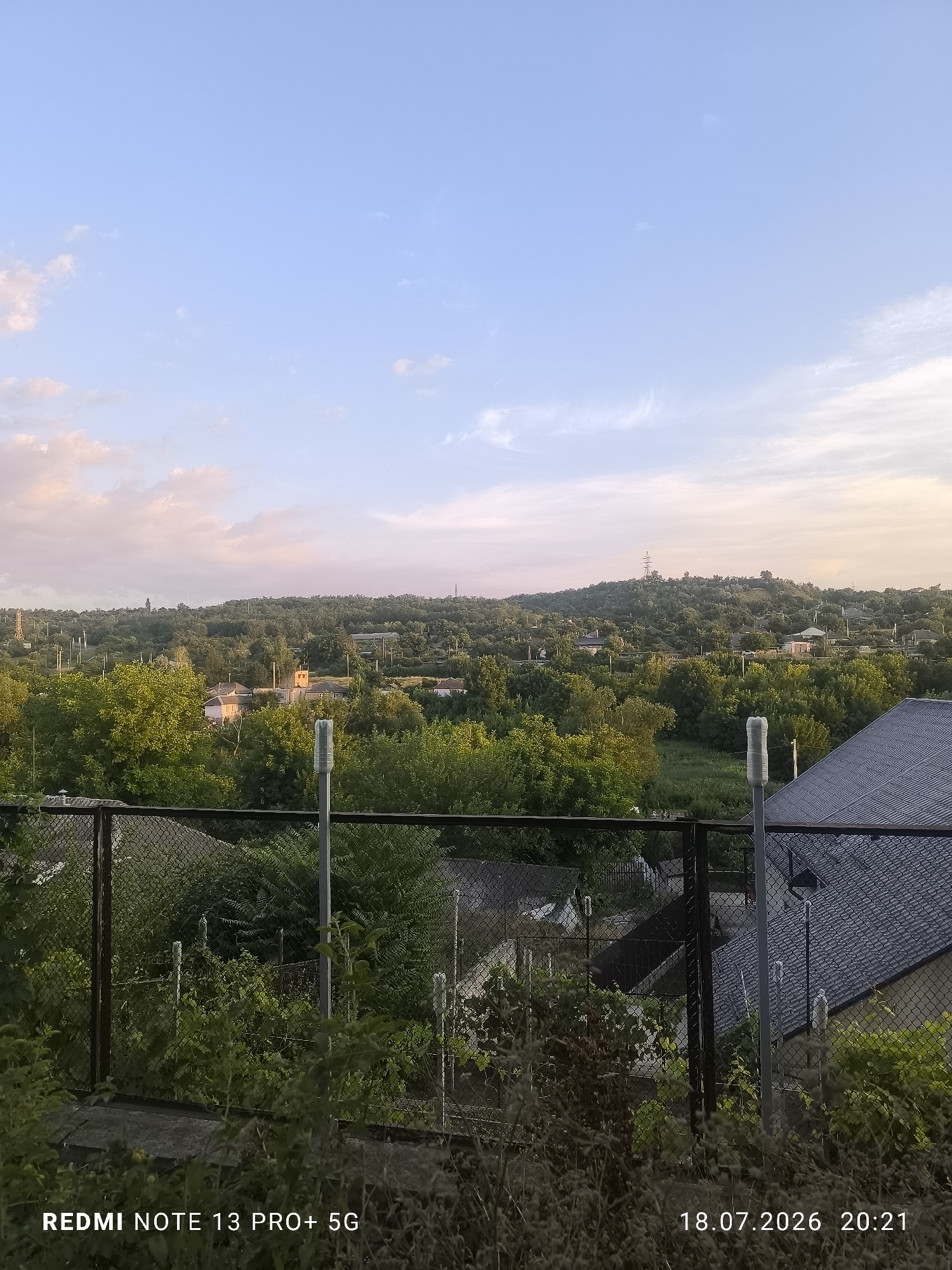 An eye-level, vertical shot captures a tranquil, panoramic view of a rural landscape at dusk. The sky, a gradient of soft blue at the top fading to a warm, pastel pink near the horizon, is adorned with wispy, scattered clouds. Below, a lush, green, tree-covered hillside dominates the middle ground, dotted with houses and illuminated by the fading sunlight. A prominent power line tower stands atop the hill. In the foreground, a dark chain-link fence with metal posts separates the viewer from a patch of overgrown greenery and the dark, shingled roof of a house. The lower portion of the image displays a timestamp: "18.07.2026 20:21" and "REDMI NOTE 13 PRO+ 5G."