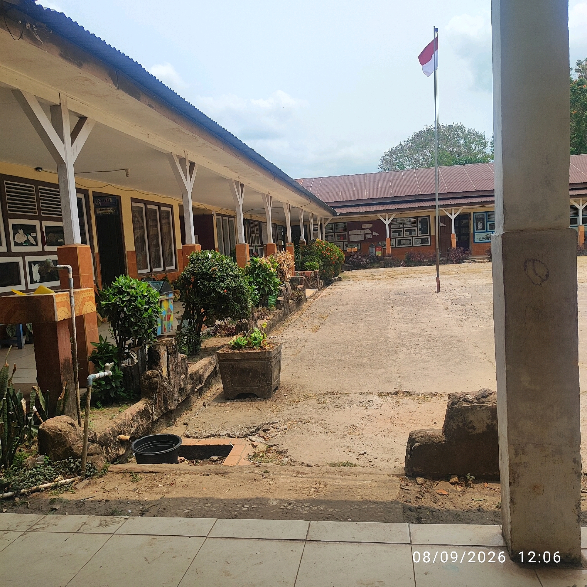 The image shows the exterior of a school building in an Indonesian city. The building features a long, covered walkway with supporting pillars and a corrugated metal roof. Along the walkway are several classrooms with windows and what appear to be displays on the walls. In front of the building, there is a courtyard with a paved area and some landscaping, including potted plants and small trees. A flagpole with an Indonesian flag is visible in the background. The scene appears to be taken during daylight hours with a cloudy sky. A timestamp is visible in the lower right corner of the image: "08/09/2026 12:06".