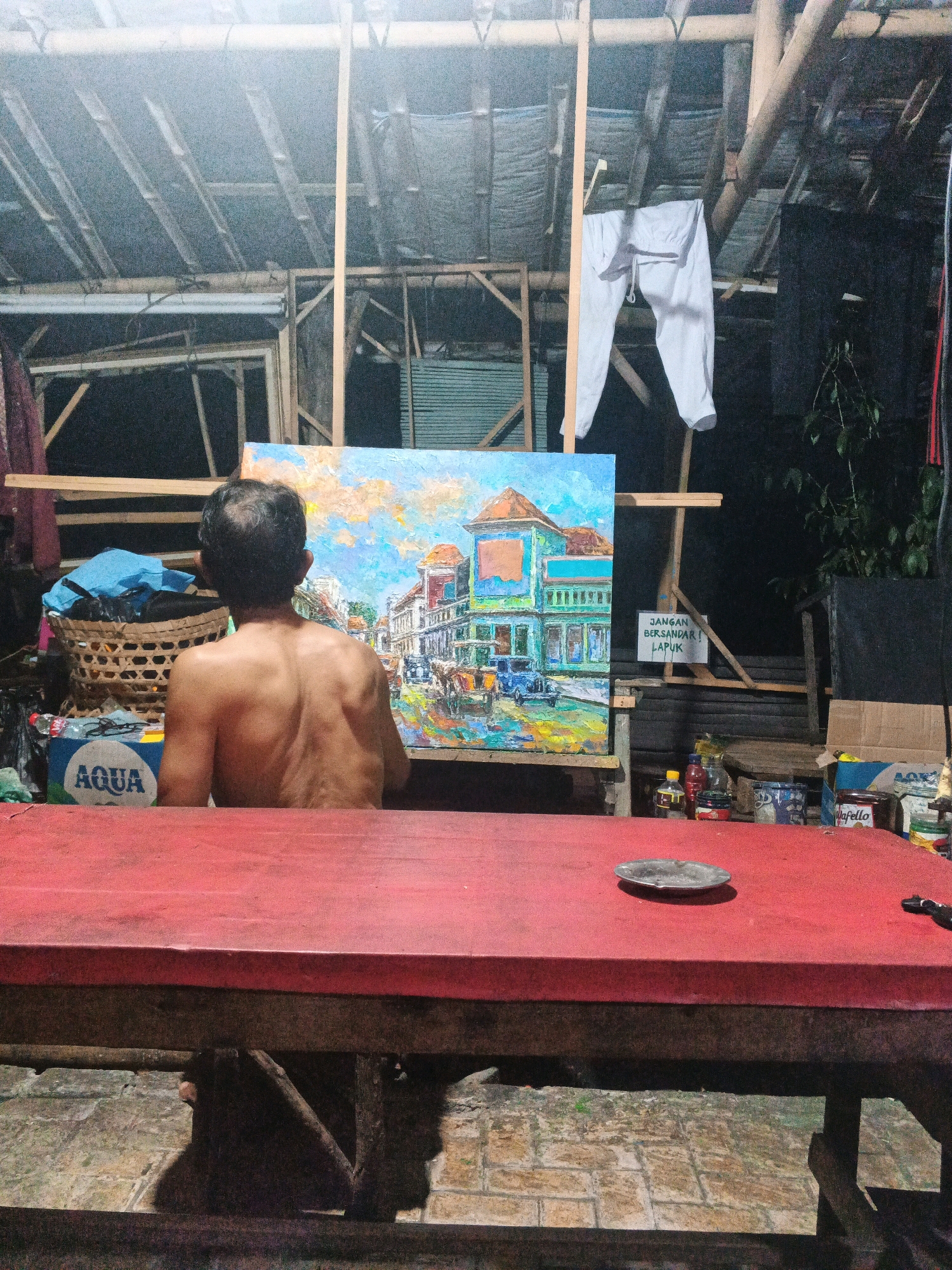 An over-the-shoulder shot shows a man from the back, shirtless, painting an outdoor scene of a street with buildings and a blue car. He is standing in what appears to be a workshop or open-air covered space with a bamboo ceiling. To the left of the painting, a woven basket contains a blue bag. A red table occupies the foreground, with an ashtray on its surface. Various bottles and cans are visible on a shelf behind the painting. A white sign with red lettering is attached to a wooden easel, reading "JANGAN BERMAIN! LAPUK!" which translates to "DO NOT PLAY! ROTTEN!". The overall setting is rustic and appears to be in a humid climate, possibly Ungaran, Indonesia, given the architectural style of the painting and the lush vegetation visible in the background, suggesting a tropical or subtropical environment.