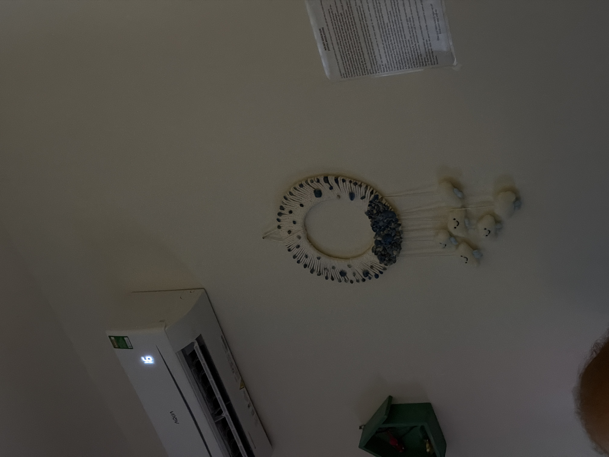 A tilted photograph captures an indoor scene, likely a wall or ceiling, in Ninh Chu Ward, Vietnam. On the left side of the frame, a white AQUA air conditioner unit is mounted, featuring a digital display showing the number "16" in blue light, and a green energy efficiency label.
Centrally positioned is a decorative, circular wall hanging made of white string or yarn, embellished with blue and white beads or small stones. Multiple strands hang from this circle, each with a small, white, plush figure attached, resembling a sheep or cloud with simple facial features.
In the upper part of the image, a piece of paper with dense printed text is affixed to the surface. While most of the text is unreadable due to distance and angle, faint words like "NEVER LEAVE US" and "KEEP YOUR O" are partially discernible at the top. To the lower right, a small, open, dark green box or shelf is partially visible, containing indistinct items. A patch of what appears to be a person's hair or head is seen in the very bottom right corner. No specific activity is taking place; the image is a static shot of the room's fixtures and decor.