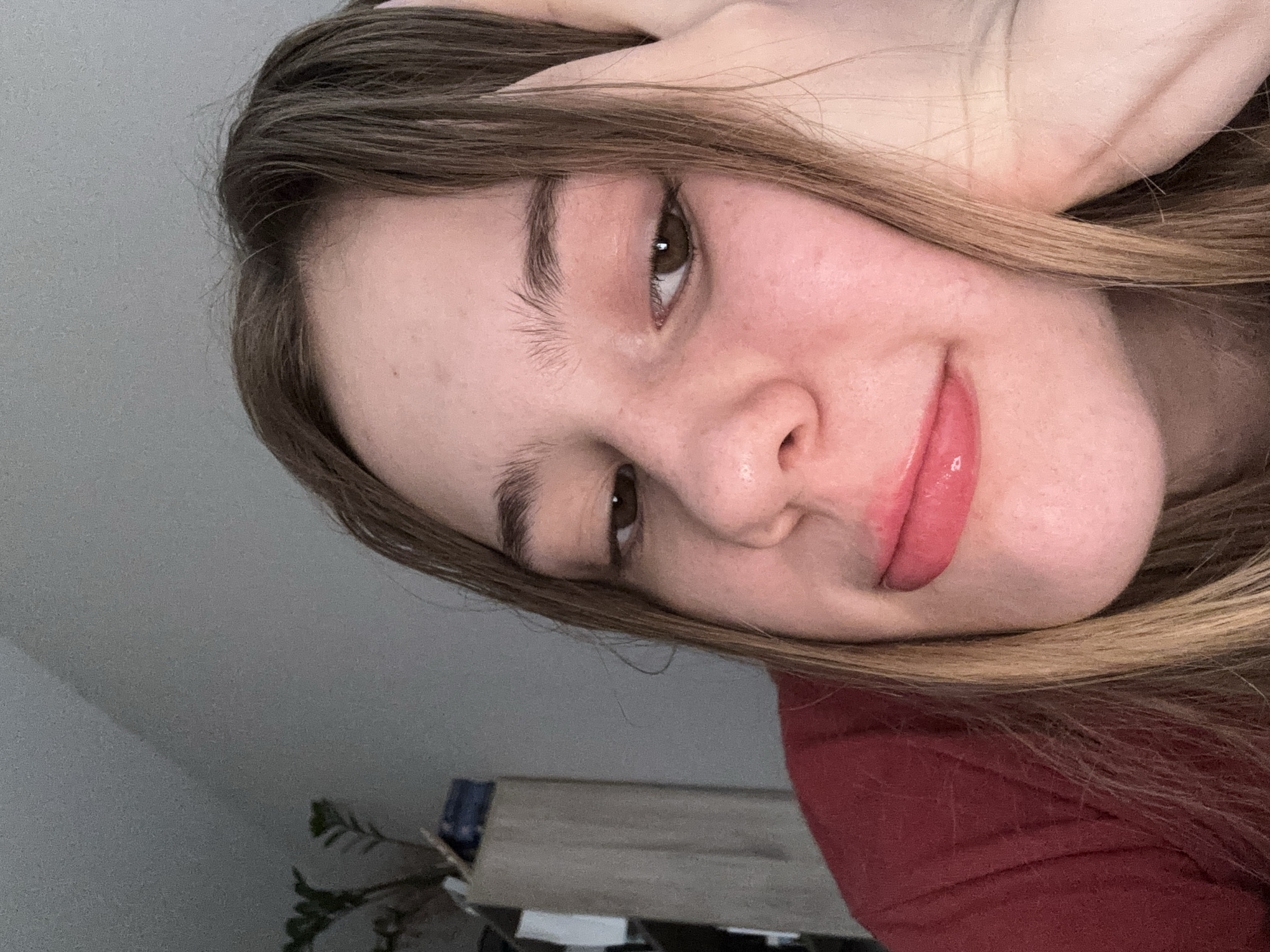 A close-up, head-and-shoulders selfie of a young person with long, light brown hair and brown eyes, resting their head on their right hand. They are wearing a red shirt. The background is out of focus and appears to be an indoor setting with a plain wall and a bookshelf with some items on it. The lighting is soft, and the overall mood is relaxed or contemplative.