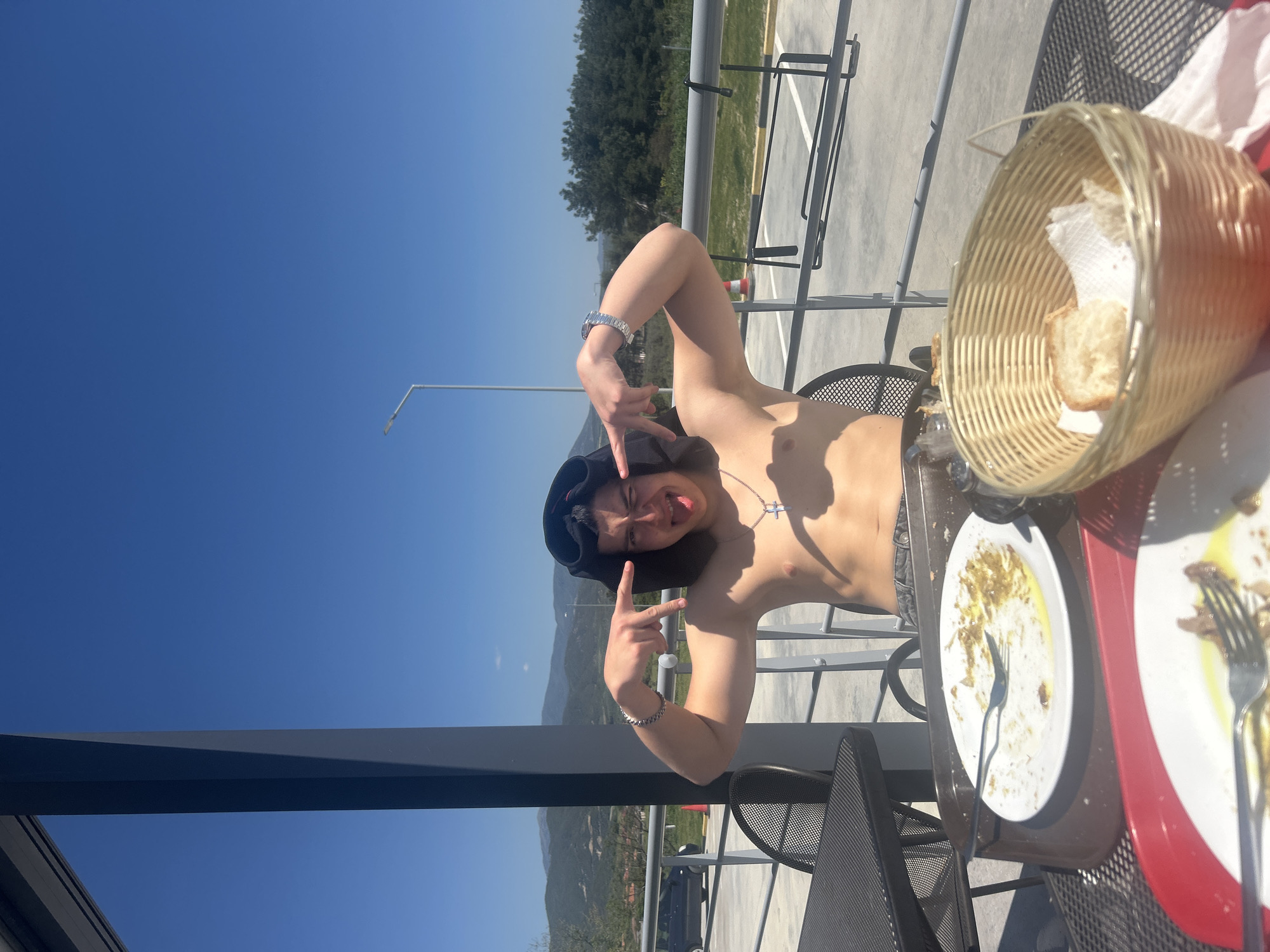 A topless young man wearing a black hat is seated at an outdoor table in Νέα Κερδύλια, Greece. He is making the "rock on" hand gesture with both hands. On the table, there are two plates with remnants of food and cutlery, and a basket of bread. The background shows a clear blue sky, trees, and a metal railing. A tall light pole is also visible.