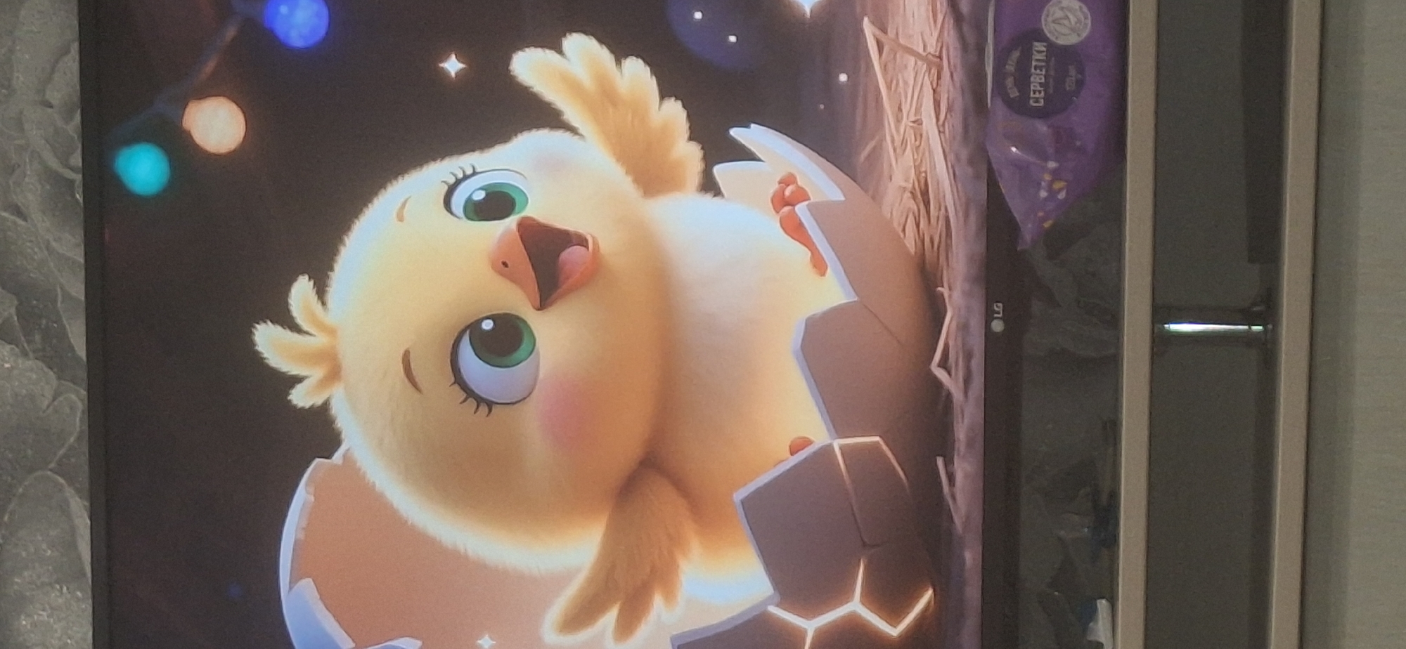 The image displays a cartoon rendering of a yellow chick emerging from a cracked eggshell. The chick has large green eyes, rosy cheeks, and its beak is open in a cheerful expression. It is situated on a bed of straw. In the background, there are blurred lights, some appearing as blue and orange spheres, and faint star-like shapes. Below the chick's perch, a portion of an LG television screen is visible, with a purple package of wet wipes in the foreground, displaying Russian text that reads "СЕРБЕТКИ" (servetki, meaning "wipes"). The environment appears to be indoors, likely a home or studio, and there is no indication of the city of Bila Tserkva, Ukraine, or any forensic details related to a crime scene.