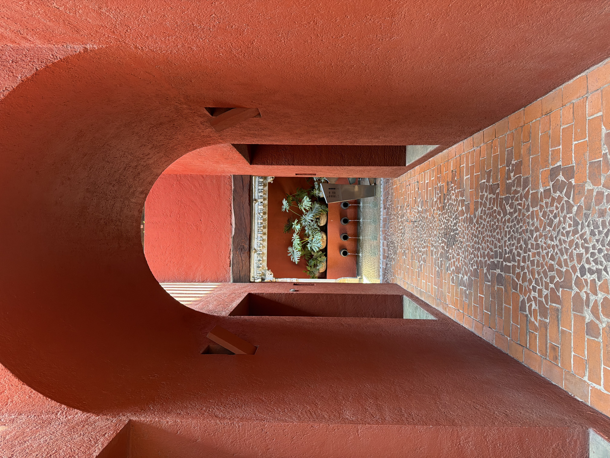 The scene depicts an interior view through a deep, arched passageway of a boutique hotel or colonial building in Puebla, Mexico, potentially the Hotel Boutique Casona Poblana, known for its distinctive red architecture.
The foreground is dominated by a robust, reddish-orange stucco archway, presenting a textured, coarse finish. The inner surfaces of the arch are in shadow, while brighter light emanates from beyond. On the inner vertical walls of the archway, two small, rectangular, angled recesses are visible, one on each side.
The pathway extending through the archway and into the brighter area is constructed of reddish-brown brick pavers. The central portion of the path features an intricate mosaic-like pattern composed of irregularly shaped, broken brick fragments or stones, flanked by more uniformly laid rectangular bricks.
Beyond the archway, the passageway opens into a well-lit courtyard or atrium. Against the far wall, which maintains the same reddish-orange stucco, a water feature is visible. It consists of a shallow, rectangular pool from which three dark circular spouts project, actively streaming water into the pool. Above the spouts, on a raised ledge, several large, green leafy plants, possibly *Monstera deliciosa*, are arranged in round, earthy-toned pots. Above the plants, a horizontal, dark wooden beam spans the wall, beneath which an ornate, carved frieze with white and gold accents is present. To the immediate right of the water feature, a vertical metallic sign or directory structure is partially visible, displaying numerical ranges (e.g., "100-149," "150-199"), suggesting room numbers or areas within a larger establishment. The environment is devoid of any individuals or active interactions. The bright, natural light illuminates the courtyard, casting clear shadows within the archway.
