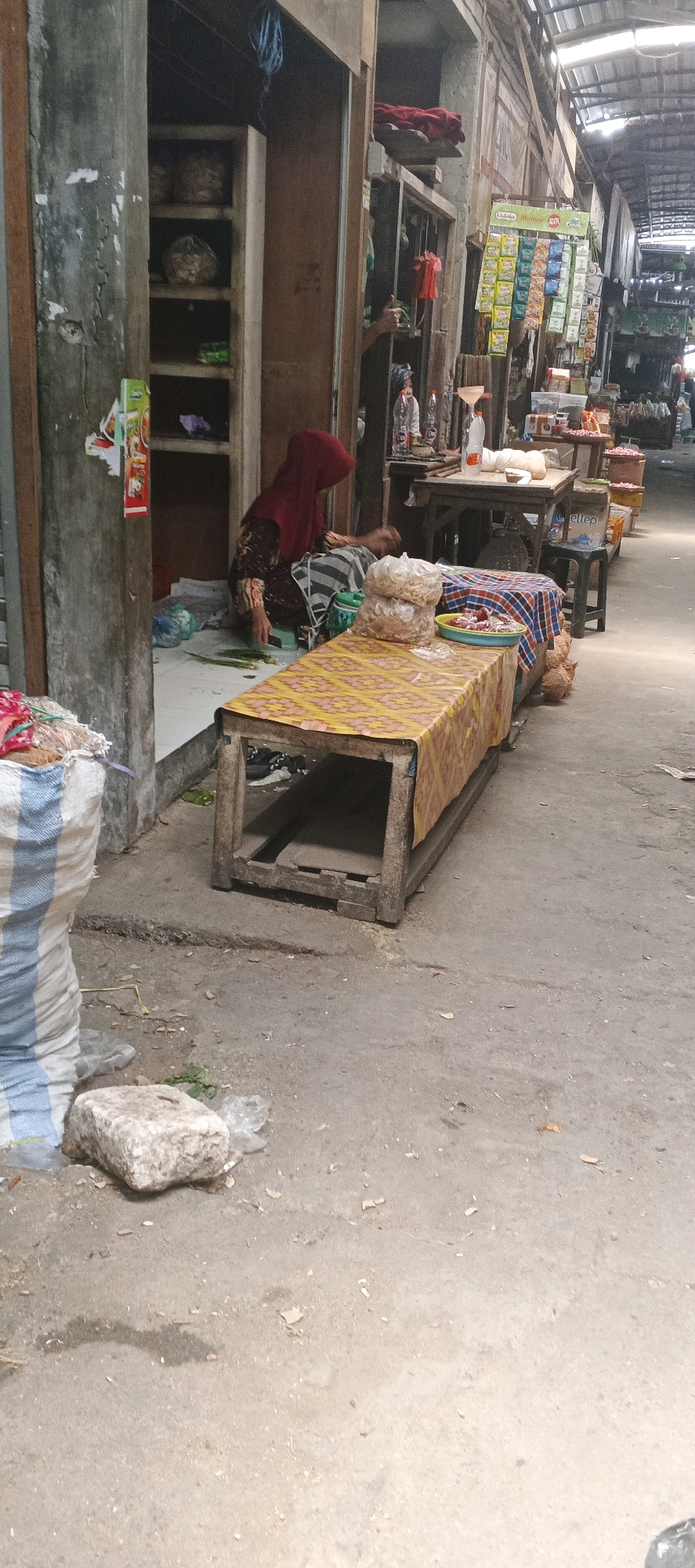 In Maduran, Indonesia, an outdoor market scene unfolds. A woman, wearing a red hijab and a brown and patterned dress, is crouched on the ground near a doorway, seemingly engaged in some activity. Before her, a wooden table, covered with a yellow and red patterned cloth, displays a mound of food items in plastic packaging. Behind the woman, shelves are visible within the doorway, holding various items. To the right of the woman, more market stalls are arranged in a line, featuring shelves stocked with goods and a variety of food items. The ground is paved with concrete, and the overall environment suggests a bustling, somewhat unorganized marketplace, with a covered roof structure overhead.