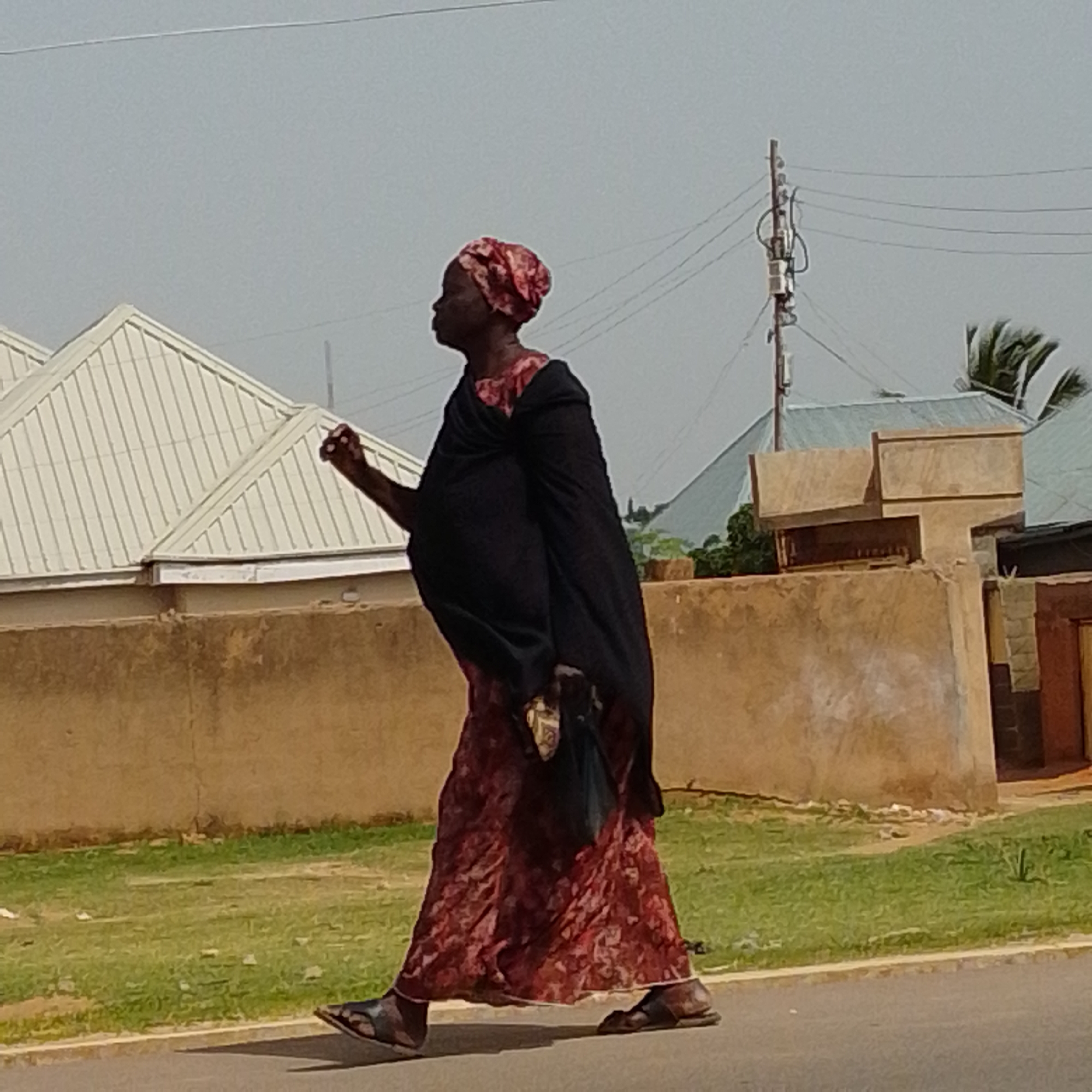 A woman is walking across a paved road, wearing a long, patterned red dress and a dark shawl draped over her shoulders. She has a red and white patterned headscarf. She is carrying a black bag. She is walking from left to right in the frame. Behind her is a low, tan-colored wall, a patch of green grass, and some buildings with light-colored roofs. In the background, there are utility poles with wires, and palm fronds are visible. The sky is a hazy gray. The scene appears to be outdoors in an urban or semi-urban environment. This image could be of a street in Gombe, Nigeria.