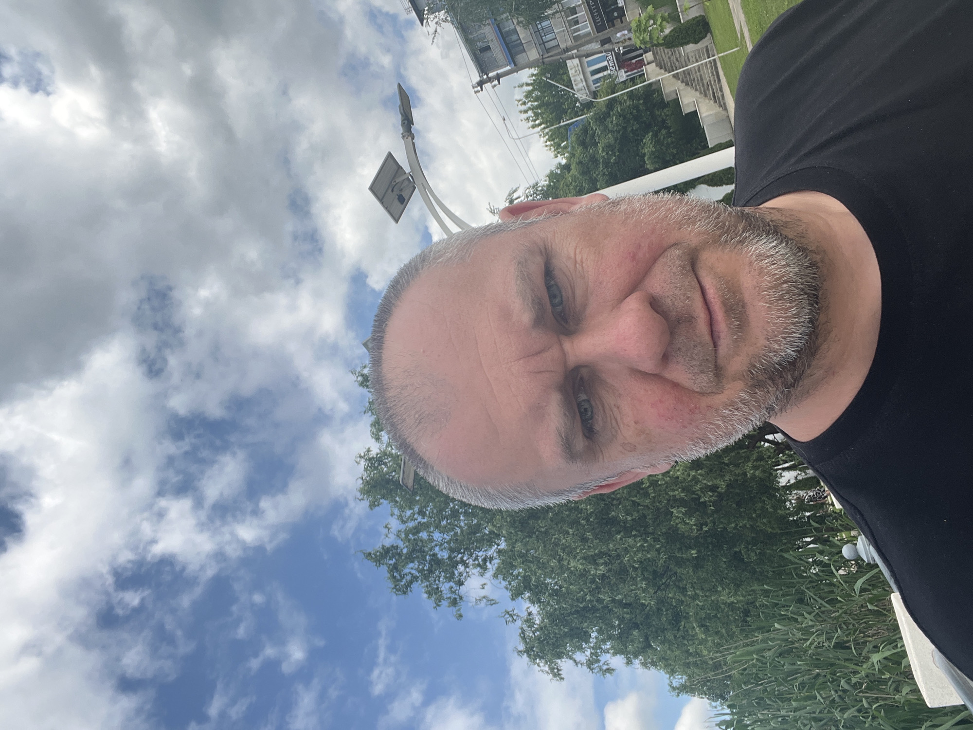 A selfie portrait taken outdoors with the subject, a middle-aged male with a graying short haircut, facing the camera and tilting his head slightly downward. He is wearing a black t-shirt.
Behind the subject, the background includes: a cloudy sky with blue patches, trees, a building with Russian Cyrillic text on it, and a street light/signage structure. The environment appears to be a city park or a roadside area with greenery in Goryachiy Klyuch, Russia.