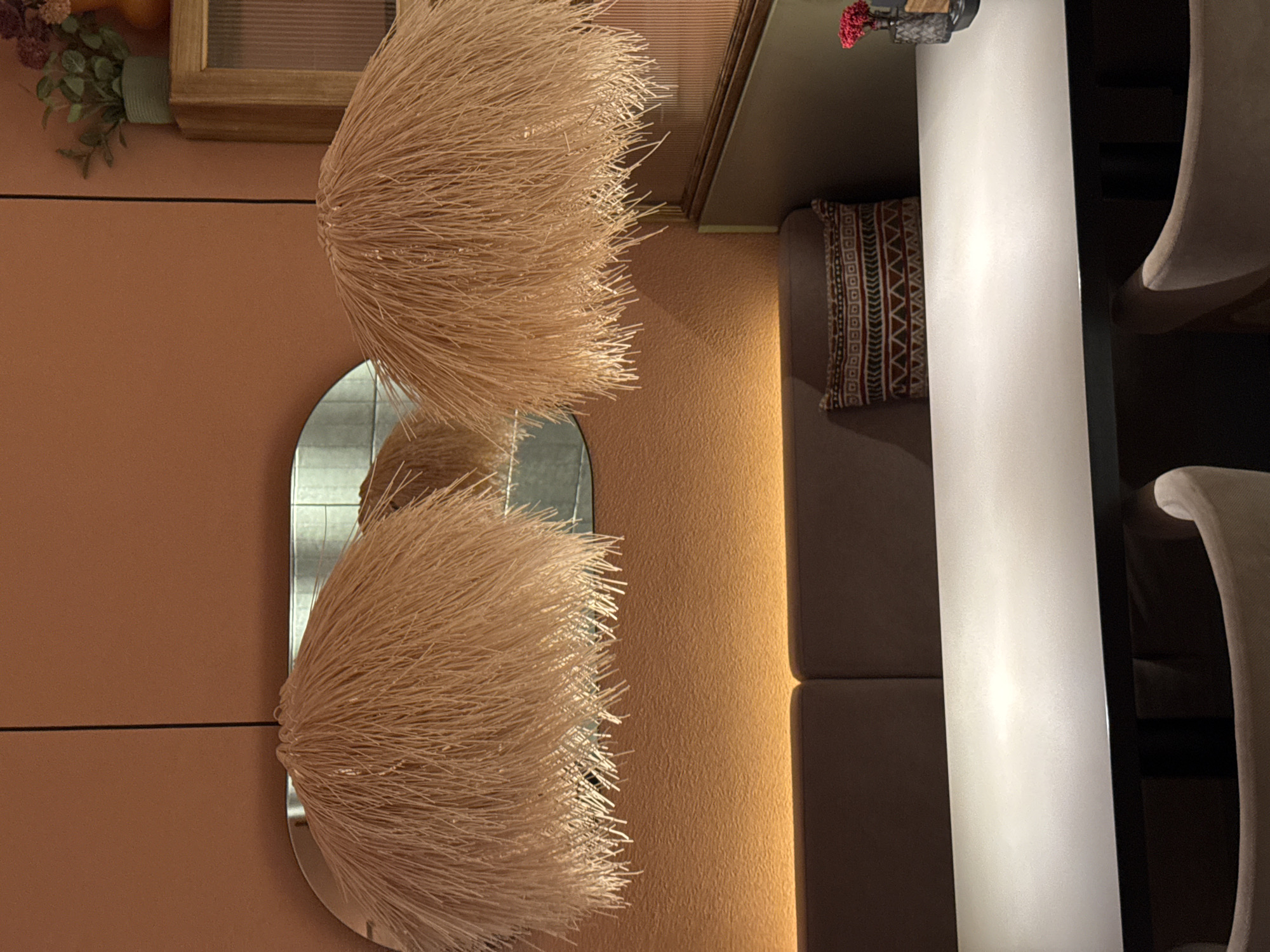 The scene depicts an interior setting in what appears to be a restaurant or cafe. On the left, there is a peach-colored wall with two decorative, straw-like spherical objects placed above and below a vertically oriented, oval-shaped mirror. Above the top spherical object, there is a framed picture or panel and a small bouquet of artificial flowers and fruits.
To the right of the wall, there is a vertical division with a muted gold tone on the left side and a seating area on the right. The seating consists of a gray, cushioned banquette with a patterned pillow. A white, shiny table runs parallel to the seating, and two dining chairs with white upholstery are visible in the background.
The details suggest a modern, stylized interior. There is no direct evidence to confirm the location in Chaikovsky, Russia, but the style and design could be indicative of a modern establishment. There are no people visible in the image.