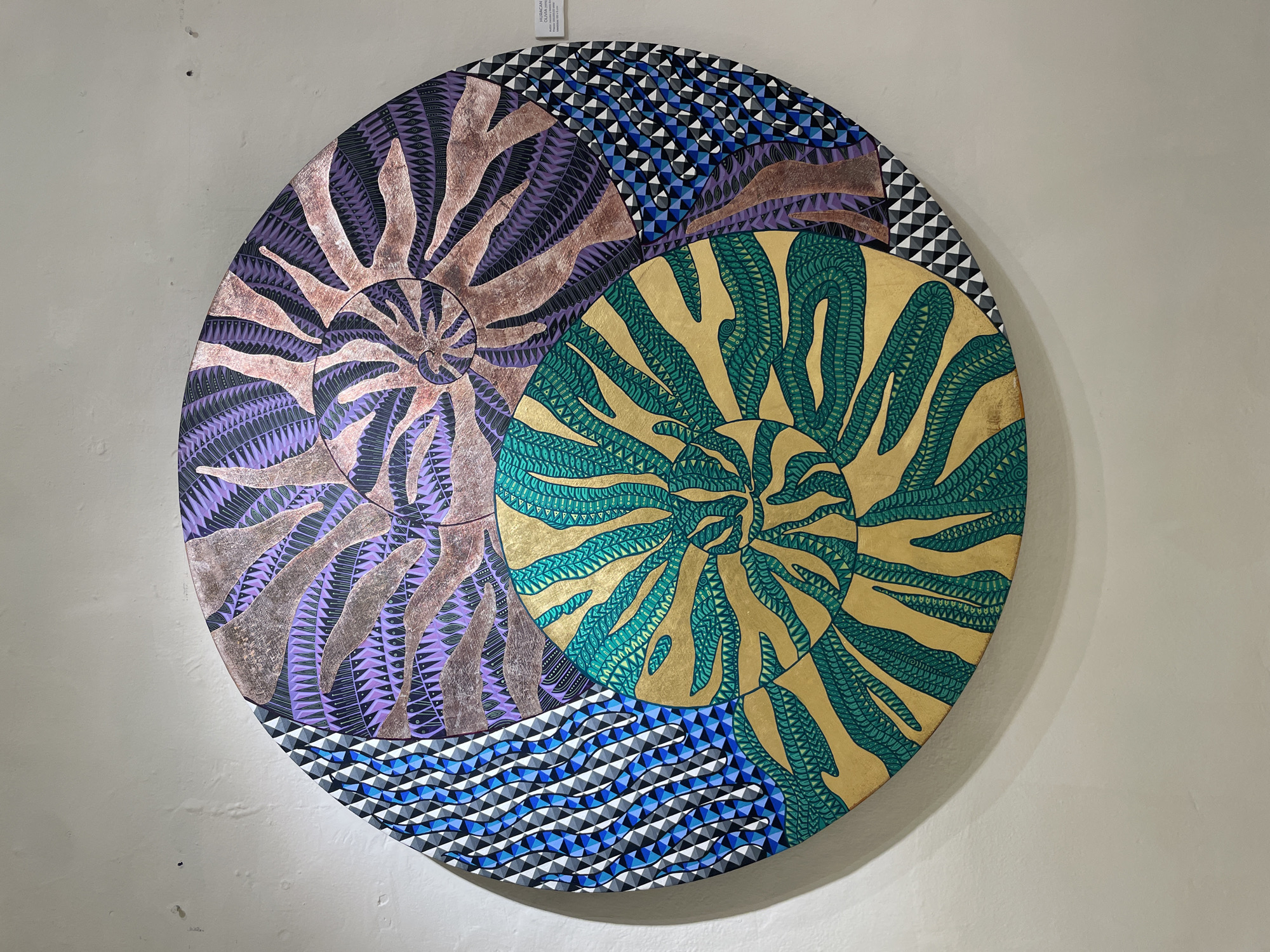 The image shows a circular piece of artwork hanging on a light-colored wall, likely in Oaxaca City, Mexico. The artwork consists of two overlapping circles, one with a purple and brown spiral design and the other with a green and gold spiral design. The remaining space around the circles is filled with a geometric pattern of blue, white, and black triangles. A small white tag is visible at the top of the artwork. The wall is plain and slightly textured.