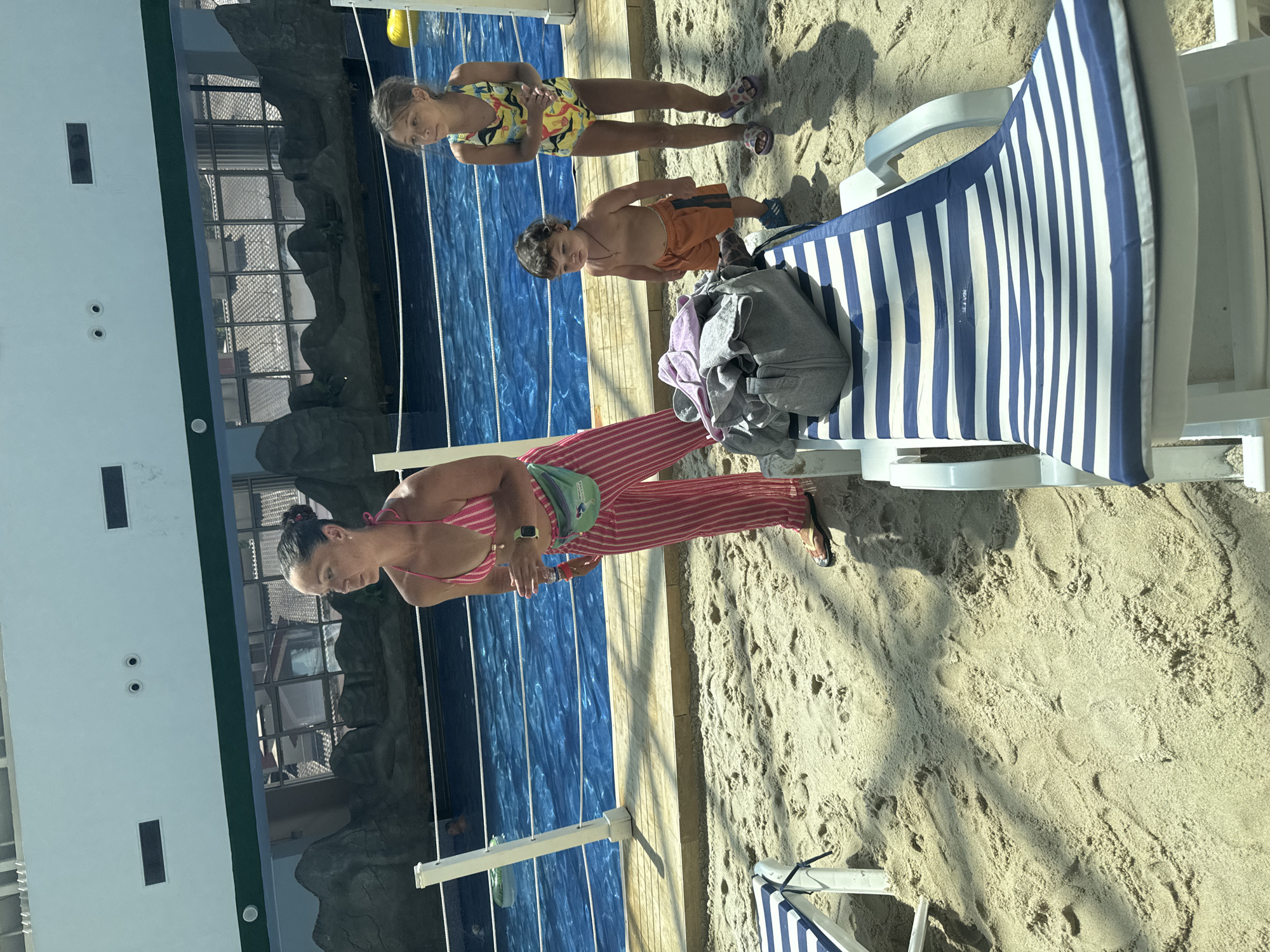 A woman in a pink striped bikini and matching pants is holding a bottle and looking down as she stands on a sandy beach next to a swimming pool. She has a green fanny pack on her waist. Two children, a girl in a yellow and blue patterned swimsuit and a boy in orange swim trunks, stand by the pool. The girl is looking at the camera, and the boy is looking forward. The pool is blue and appears to be indoors, with a large white building with windows on one side. The beach has sand and a lounge chair with blue and white stripes. The sun appears to be casting shadows, suggesting it is daytime. The location is identified as Estosadok, Russia.