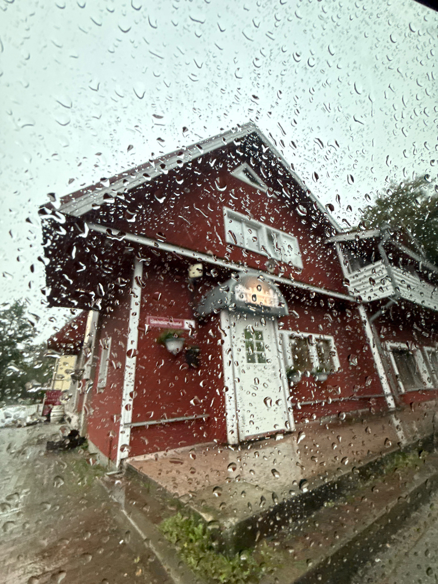 The image depicts a red building with white trim, seen through a window covered in raindrops. The building appears to be a two-story structure with a prominent gable. It has several windows, some with white frames, and a white door with a glass pane. Above the door, there is an awning and a light fixture. A sign with red text is partially visible on the side of the building, appearing to read "Sõõrumaa Hotell". A potted plant hangs near the sign. The ground in front of the building is wet and reflects the building. Trees are visible in the background. The sky is overcast, suggesting rainy weather. The perspective suggests the photograph was taken from inside a vehicle or a sheltered area looking out. The location is identified as Sõõrumaa Hotell in Tallinn, Estonia.