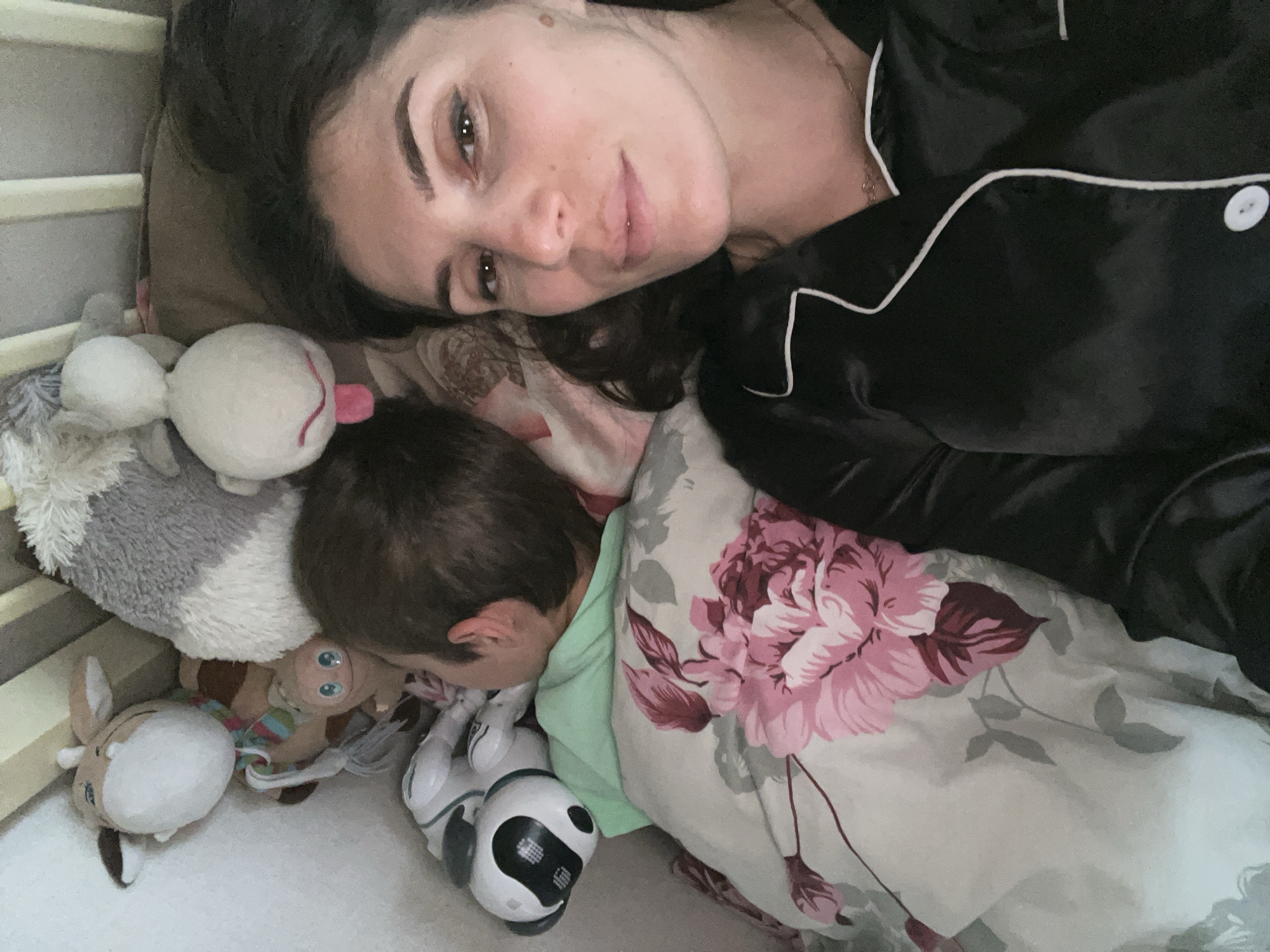 The image shows an intimate indoor scene of a woman and a young child resting together, likely in a bed or crib. The woman, positioned on her back with dark hair spread around her head, looks towards the camera with a calm expression. She is wearing black satin pajamas with white piping.
Beside her, a child lies on their side, back to the camera, wearing a light green top. Their face is not fully visible, suggesting they are sleeping or peacefully resting. Several plush toys are nestled around the child, including a white round toy with a pink tongue, a grey and white fluffy animal, a brown and white deer-like figure, a small brown plush with blue eyes, and a white and black robot toy.
A wooden crib railing is partially visible on the left side of the frame, confirming the setting as either a bed adjacent to a crib or a larger family bed. The bedding features a distinct floral pattern with large pink and maroon flowers on a light-colored fabric. The overall scene conveys a sense of peace and closeness. There is no visible text in the image. The activity appears to be quiet rest or sleep. The location is contextualized as Kaspiysk, Russia.