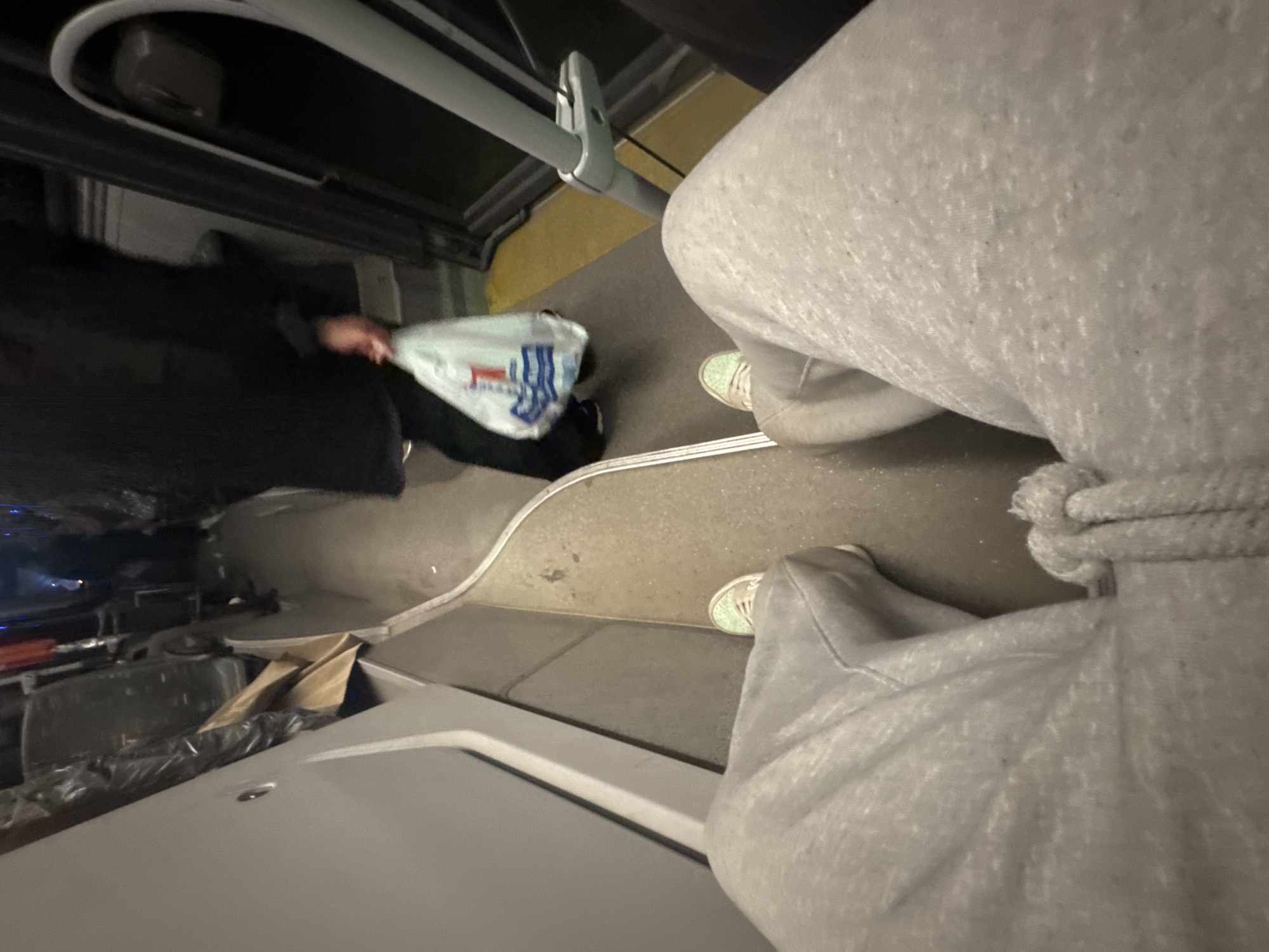 The image depicts the interior of a bus in Yerevan, Armenia. The perspective is looking down from the standing area. A person wearing gray sweatpants and green sneakers is visible. Another person in a black coat is seated, holding a white shopping bag. There is a metal safety bar running along the aisle. Bags and other items are visible near the front of the bus. The bus floor is gray.