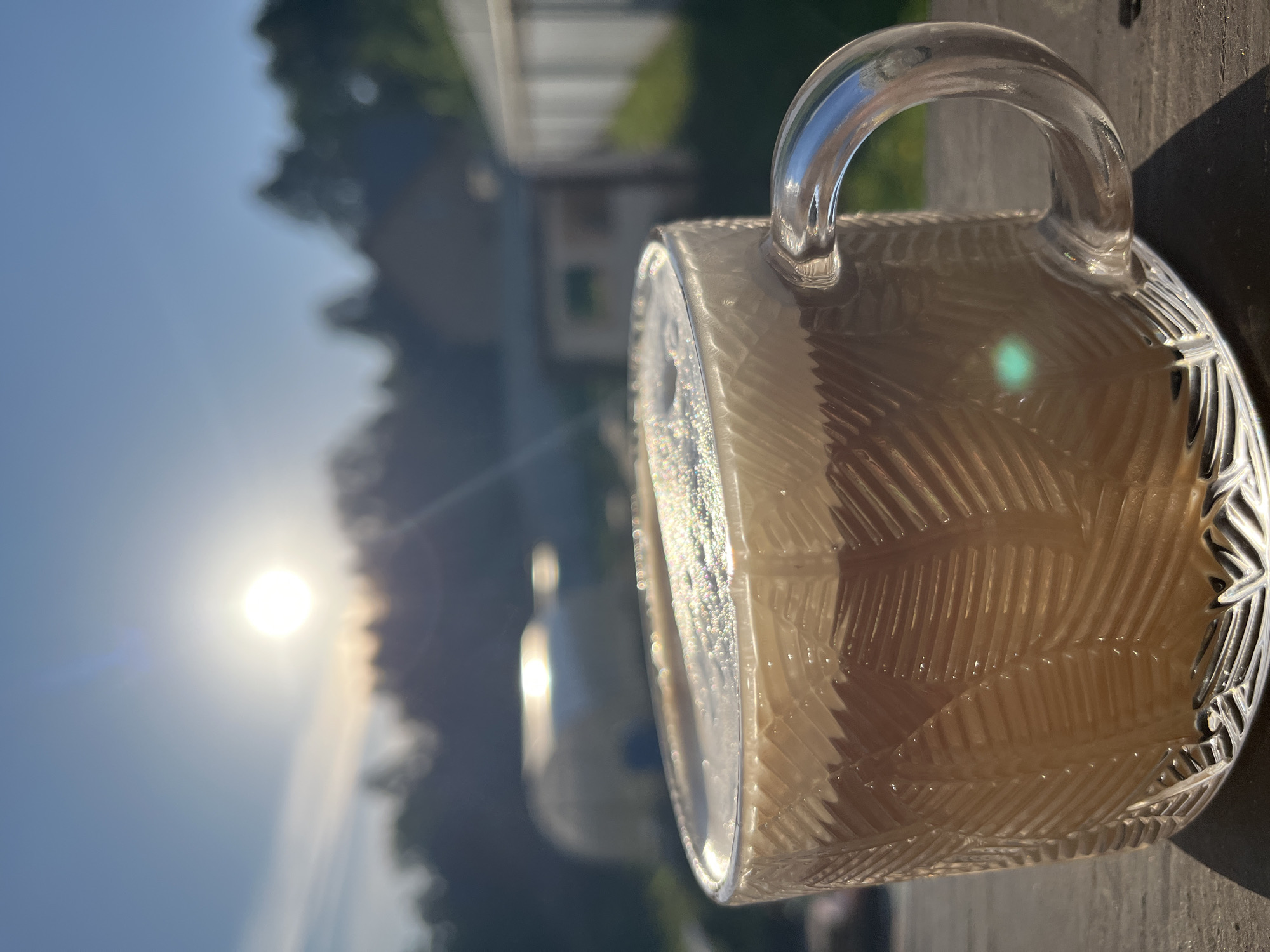 A clear glass mug containing a light-colored liquid sits on a wooden surface. Sunlight glints off the mug, and in the blurred background, there's foliage and a structure, possibly a building. The exact location within Chaikovsky, Russia, cannot be determined from this image. There are no people visible.