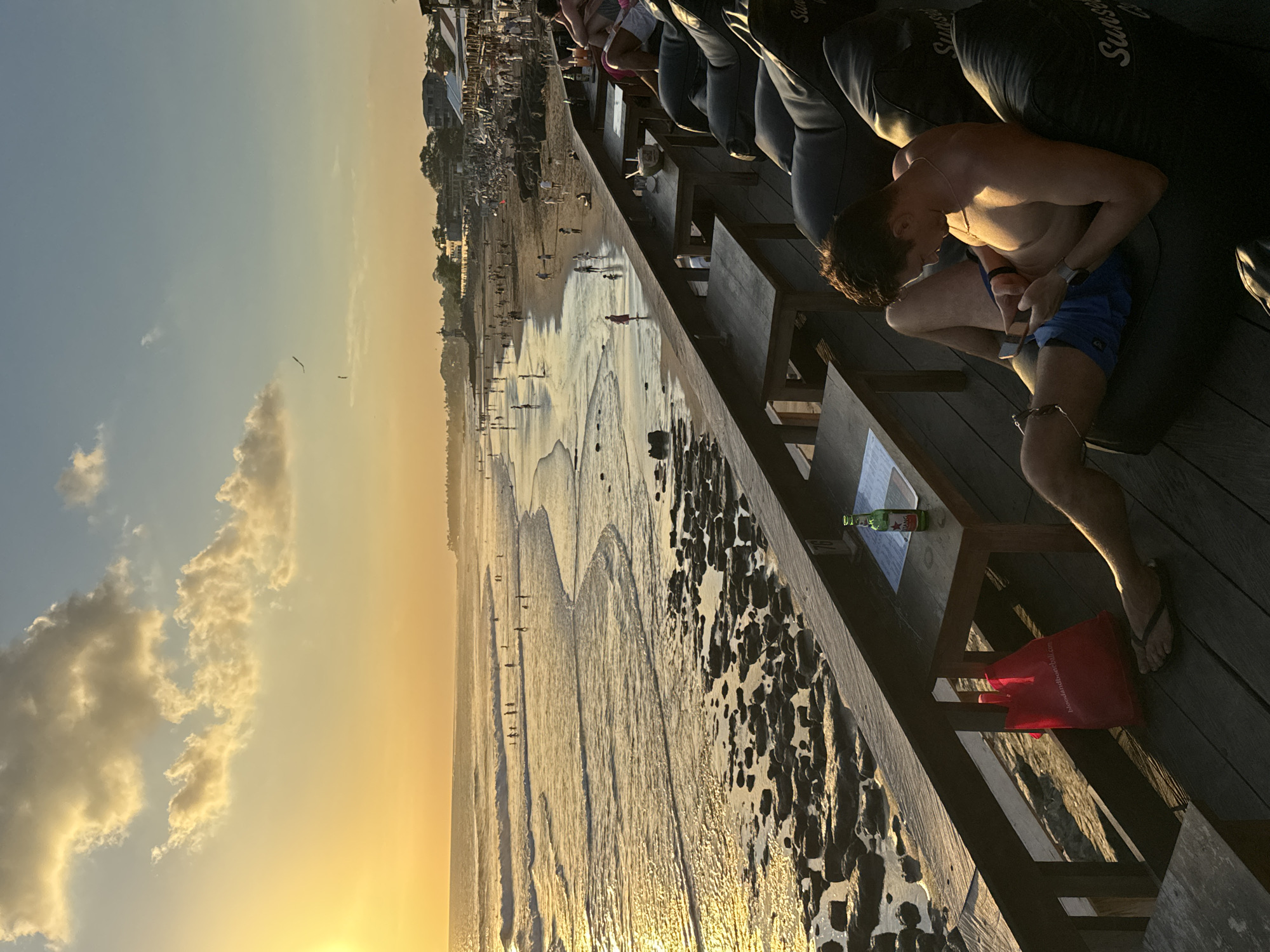 A horizontally-oriented photograph captures a scene on a beach at sunset. The location is in North Kuta, Indonesia, as indicated by the beach and the "Sunset..." text on the cushions.
In the right foreground, a shirtless man wearing blue shorts is seated on a beanbag chair, focused on his phone. Next to him on a small wooden table, a bottle of beer is visible. A red tote bag rests on the wooden deck below.
The background showcases a beach with numerous people scattered along the shoreline and buildings lined along the beach. The sky is painted with hues of orange and yellow from the setting sun, with scattered clouds enhancing the sunset ambiance. The beach is wet and reflective, mirroring the colors of the sky and creating a shimmering effect.